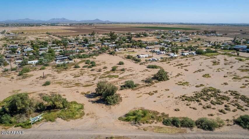 Vacant sandy parcel with scattered shrubs, neighboring homes, farmland, and distant mountains.