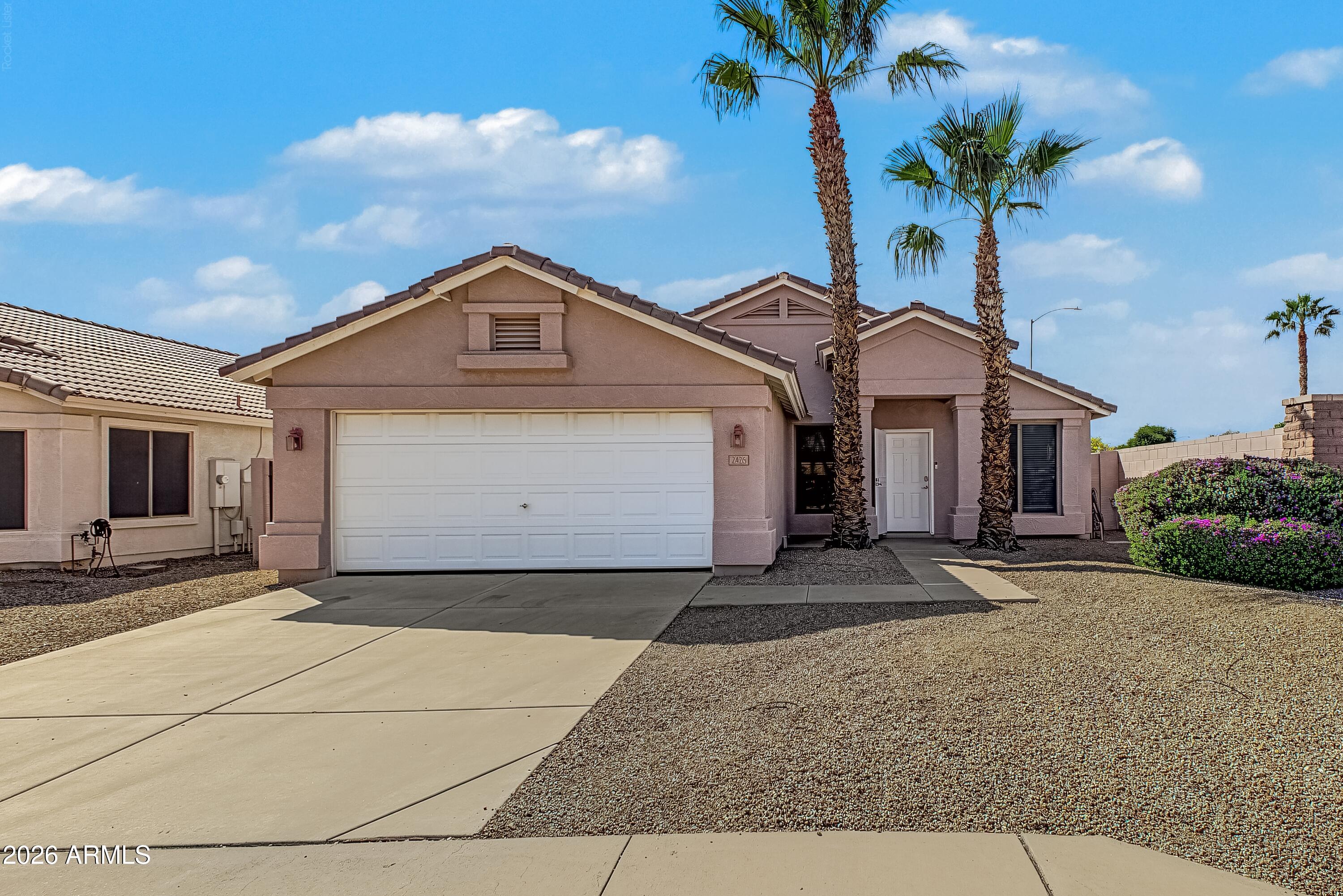 Single-story stucco home with a two-car garage, gravel yard, flowering shrubs, and tall palm trees.