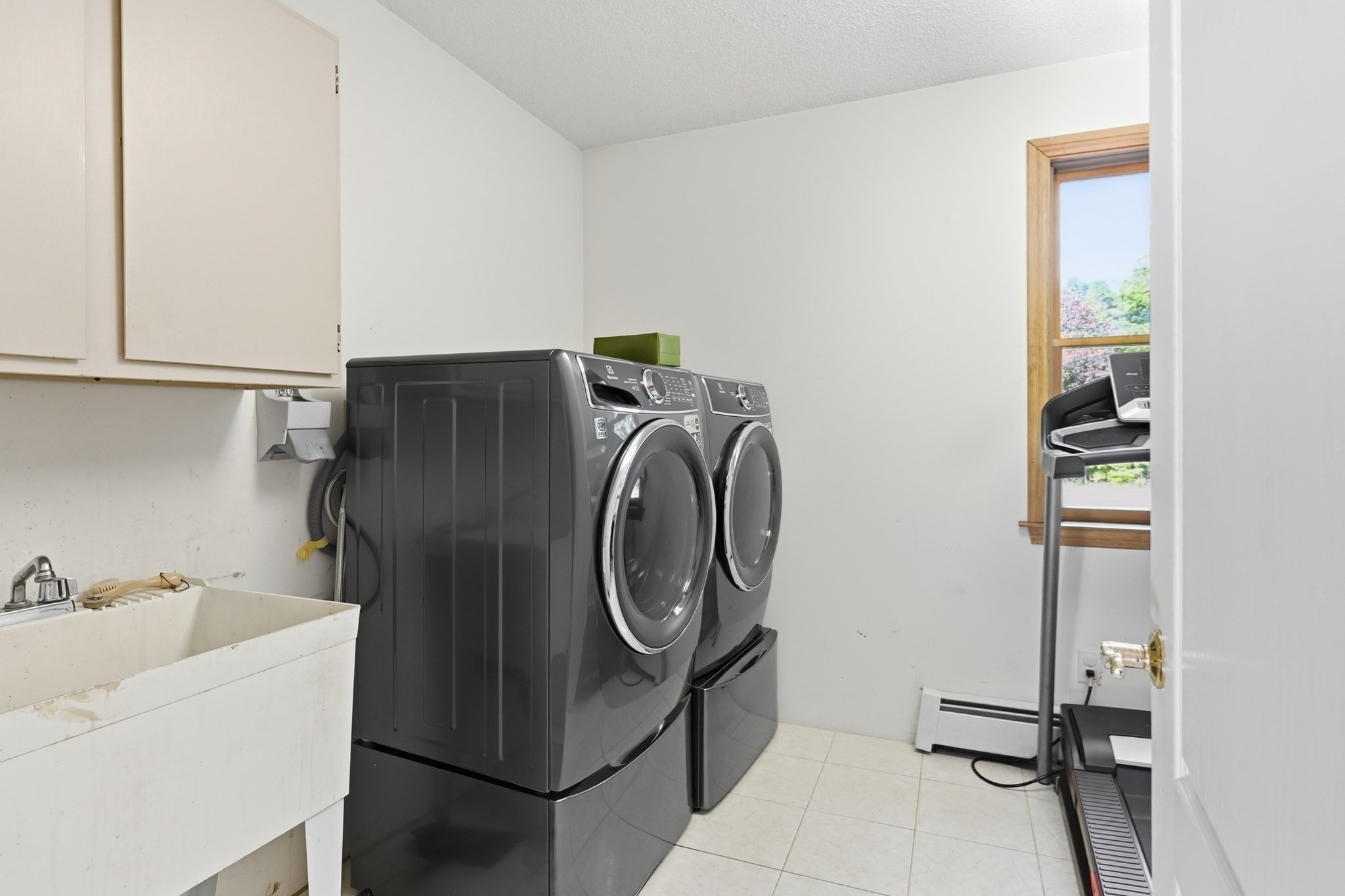 Laundry room with dark front-loading washer and dryer, utility sink, and treadmill by a window.