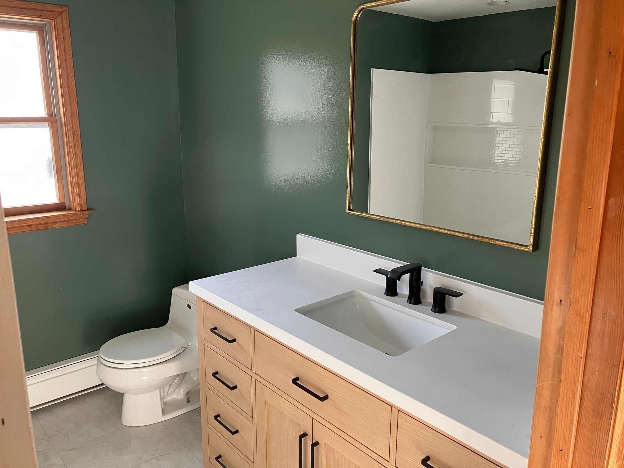 Bathroom with deep green walls, a light wood vanity, black faucet, and wood-trimmed window.