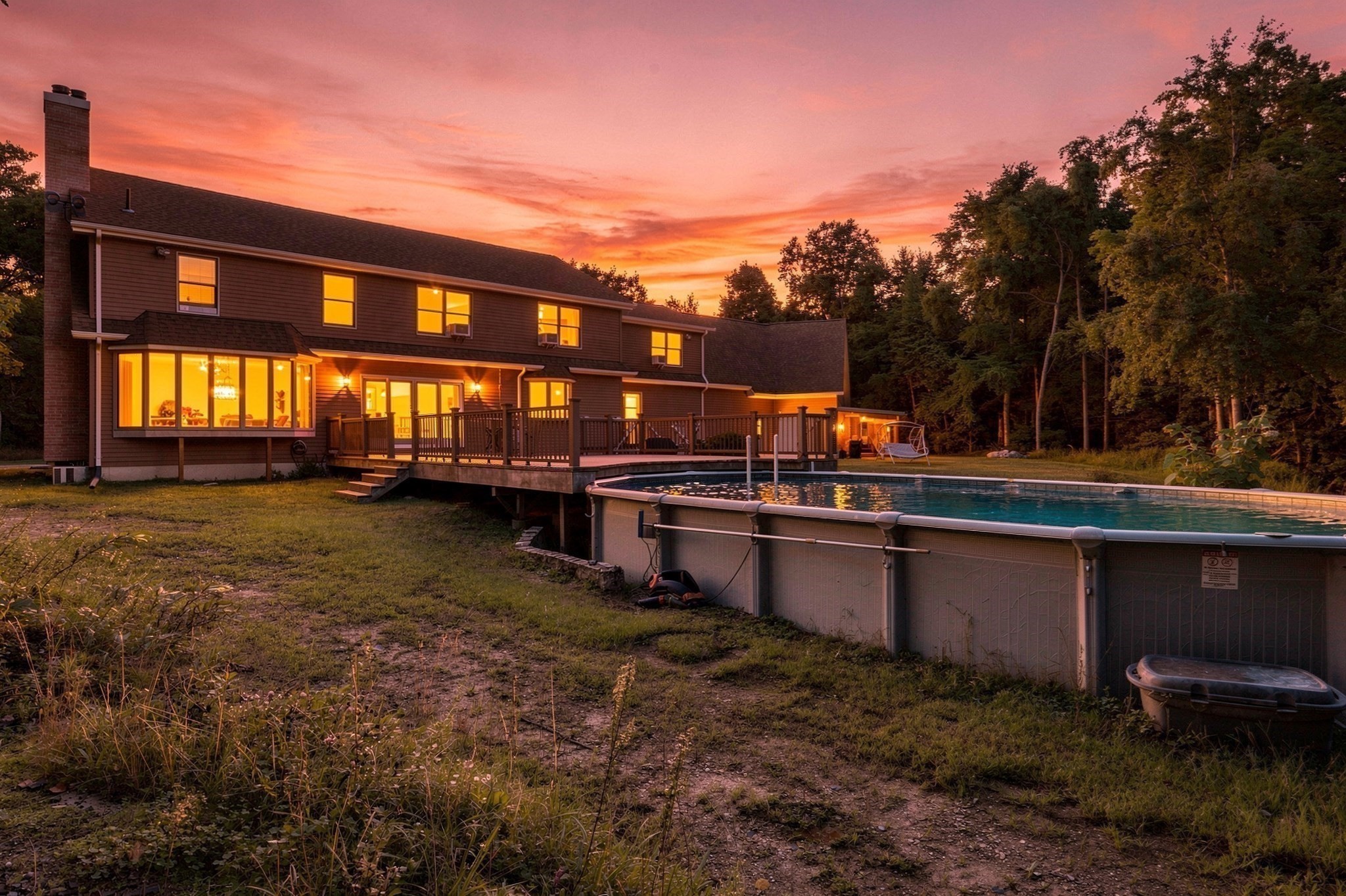 Two-story home with a lit deck and above-ground pool beneath a pink-orange dusk sky.