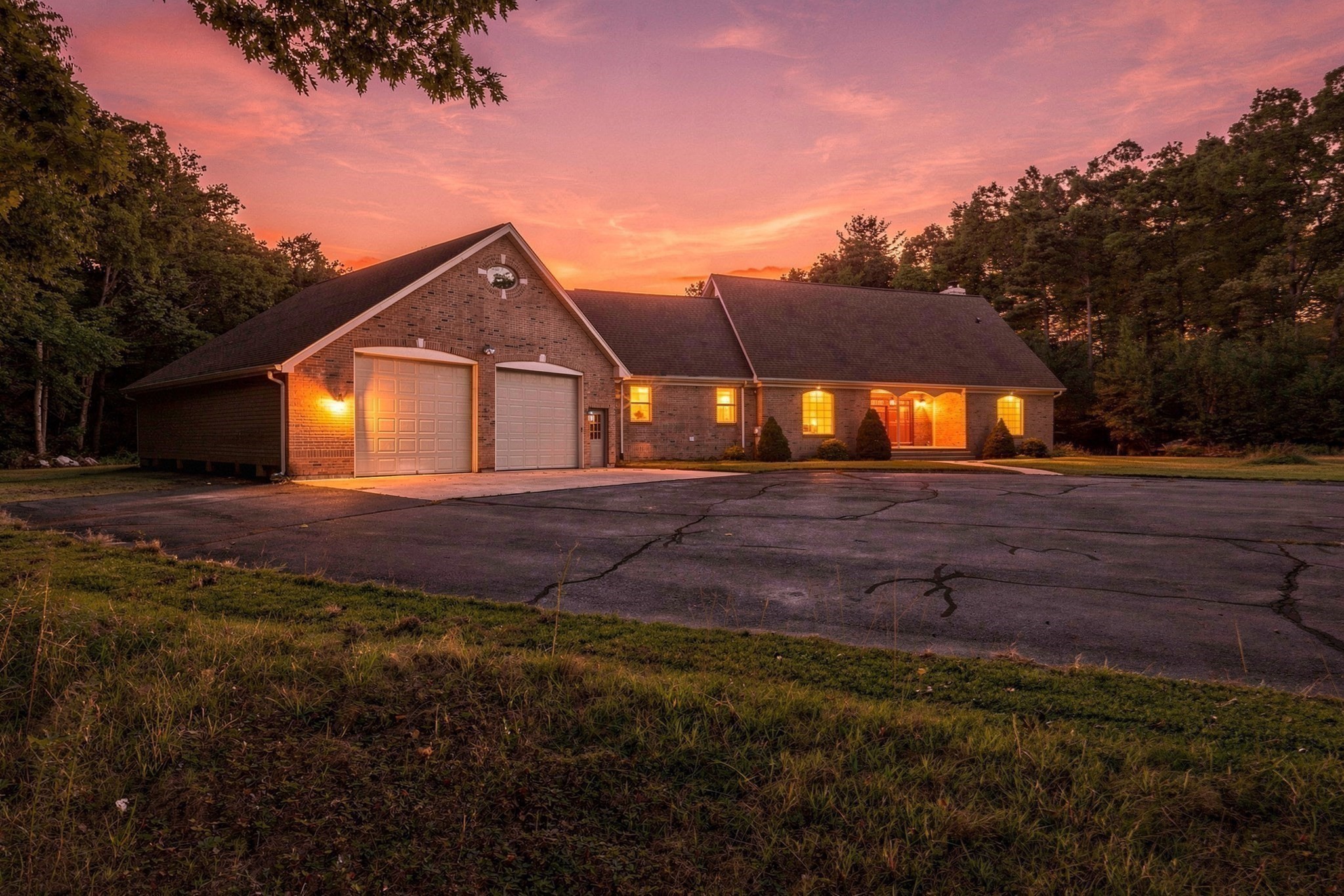 Brick house with two garage doors and warmly lit windows among trees at sunset.