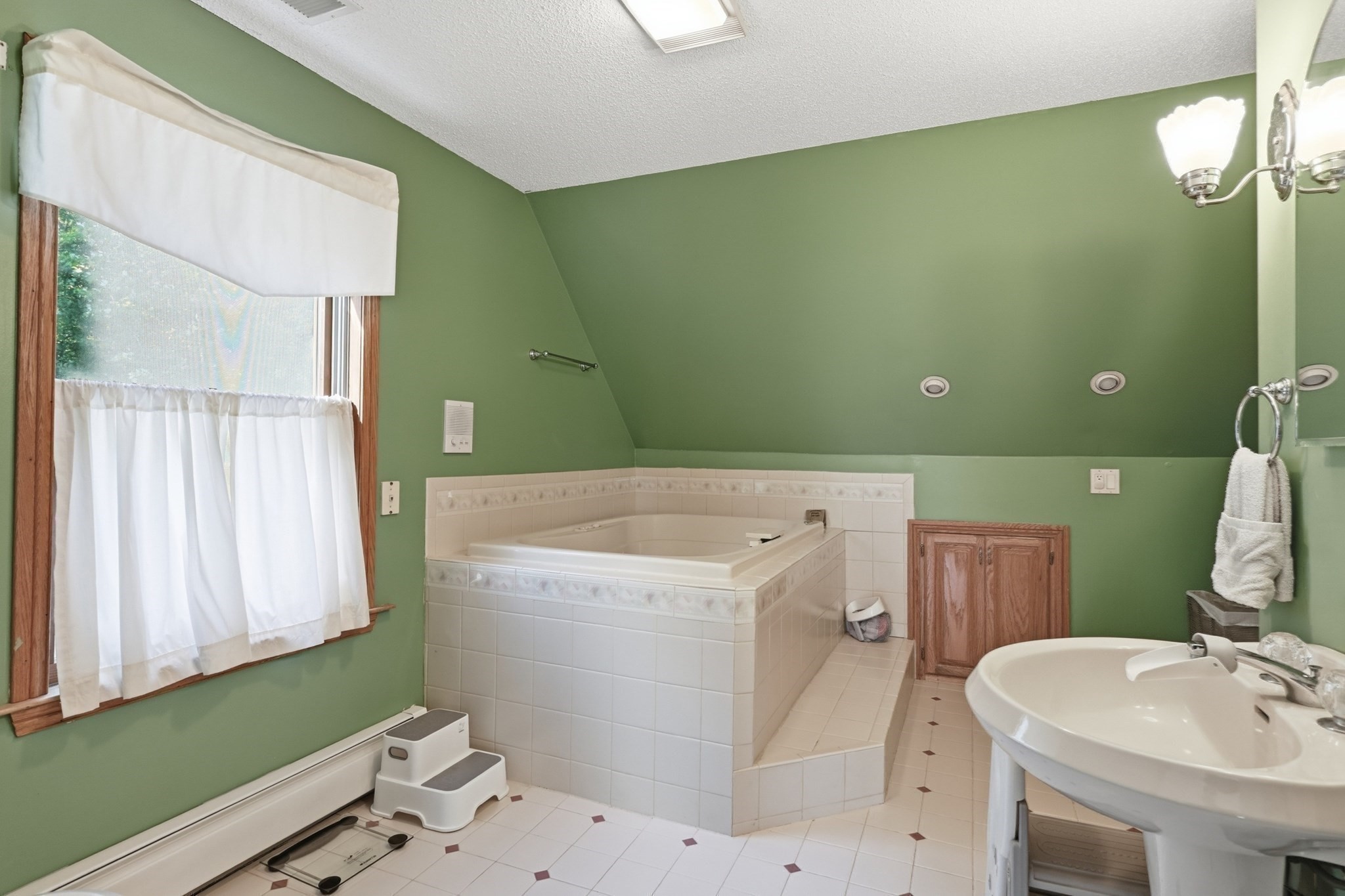 Bathroom with green walls, raised tiled soaking tub, pedestal sink, and curtained window.