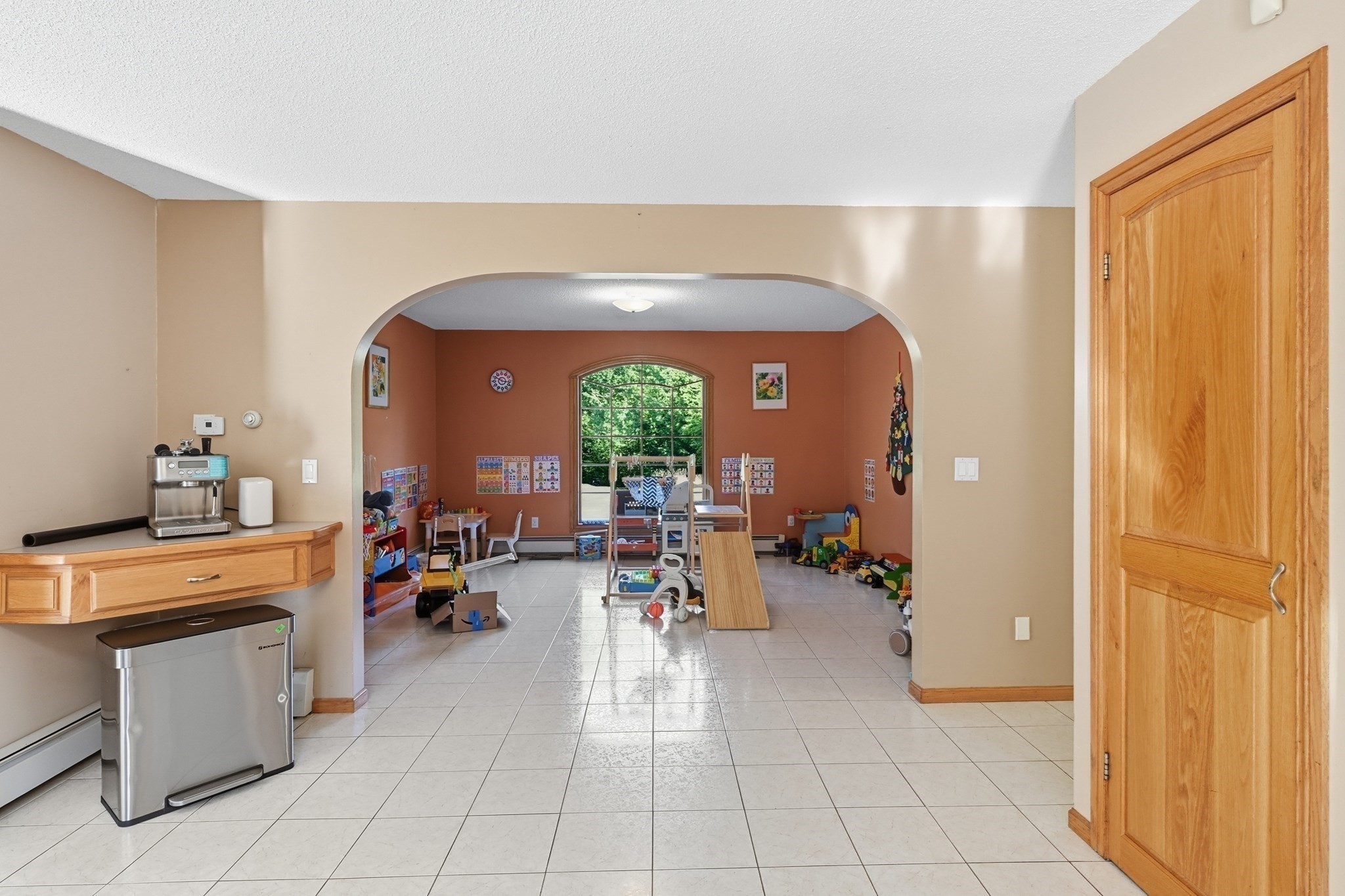 Tiled interior opens through an archway to a children's playroom with toys and a large arched window.
