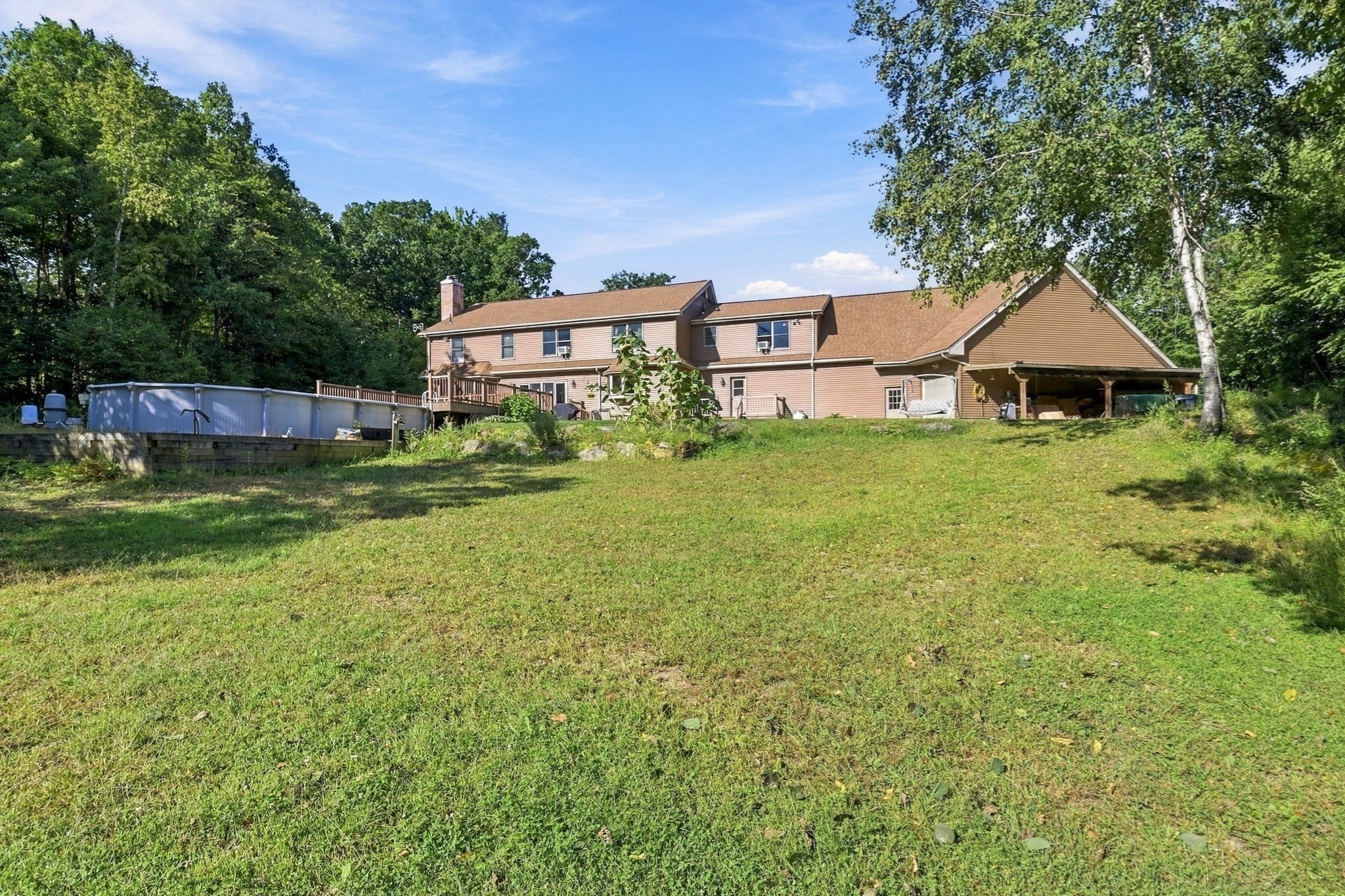 Rear of a two-story home with a raised pool, deck, covered carport, and broad lawn.
