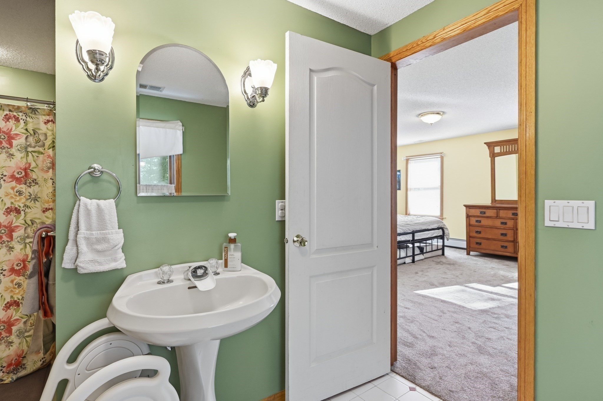 Green bathroom with pedestal sink and floral shower curtain opening onto a carpeted bedroom.