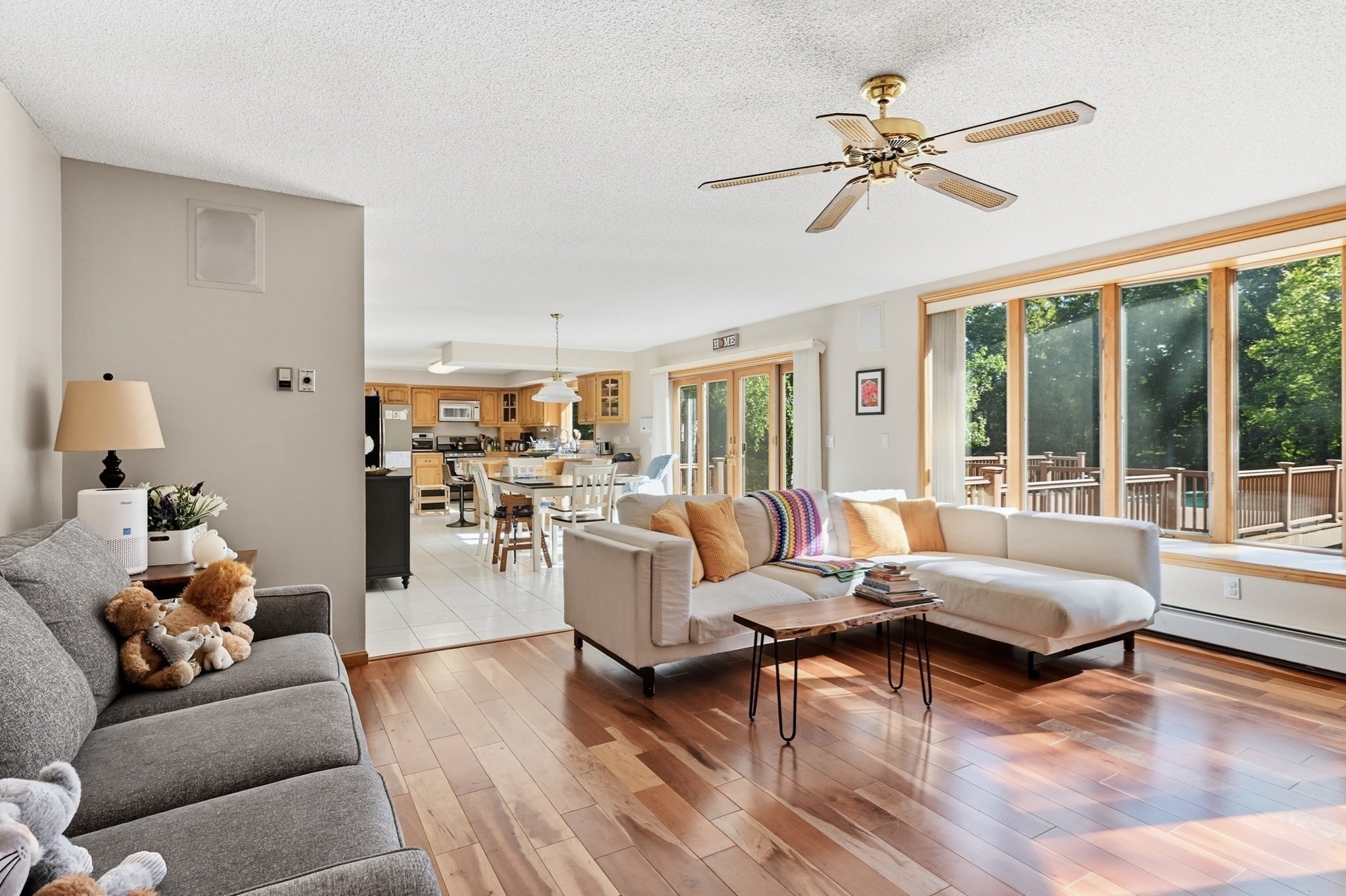 Open-plan living room with a large sectional, hardwood floors, and wide windows overlooking a deck and trees.