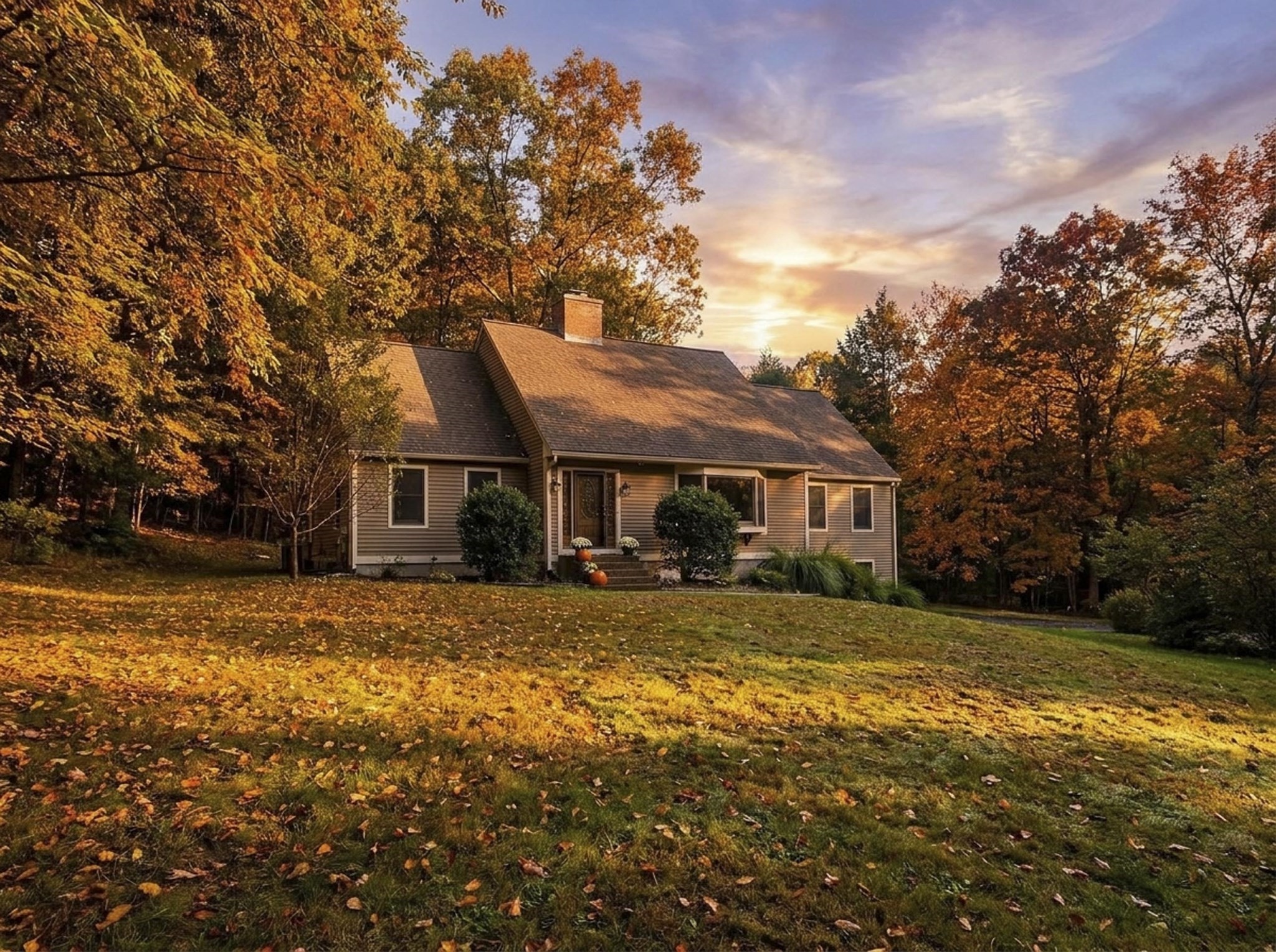 House exterior on a leaf-covered lawn, framed by autumn trees at sunset.