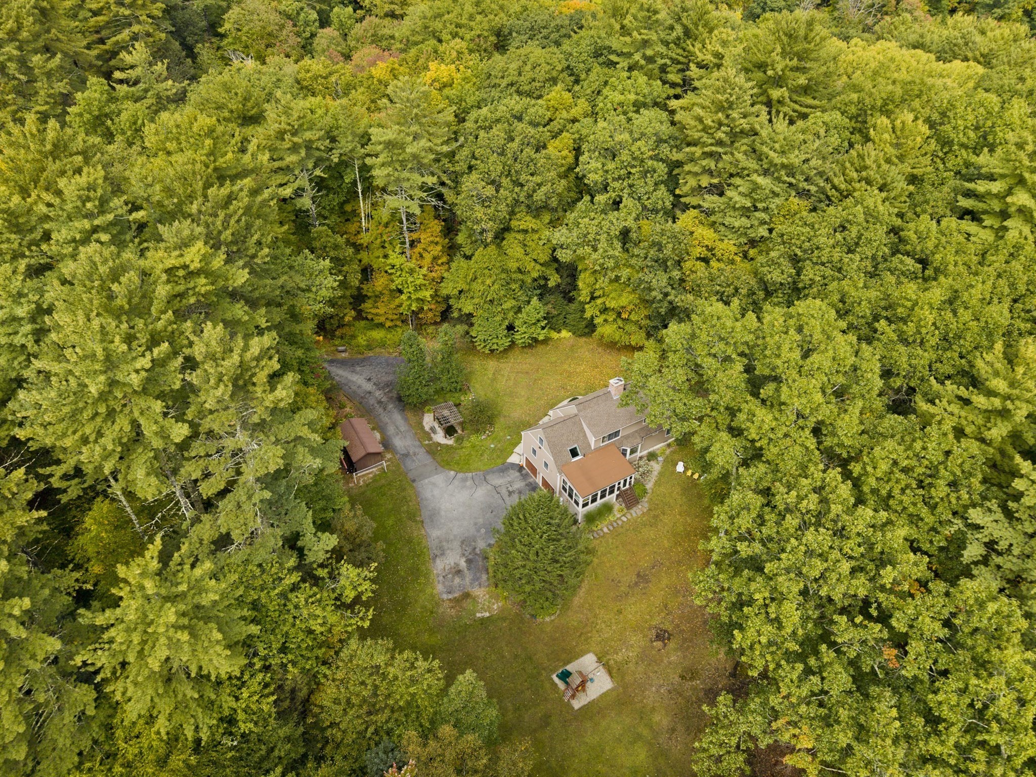 Aerial view of a house surrounded by trees, with a long driveway, detached shed, and open lawn.