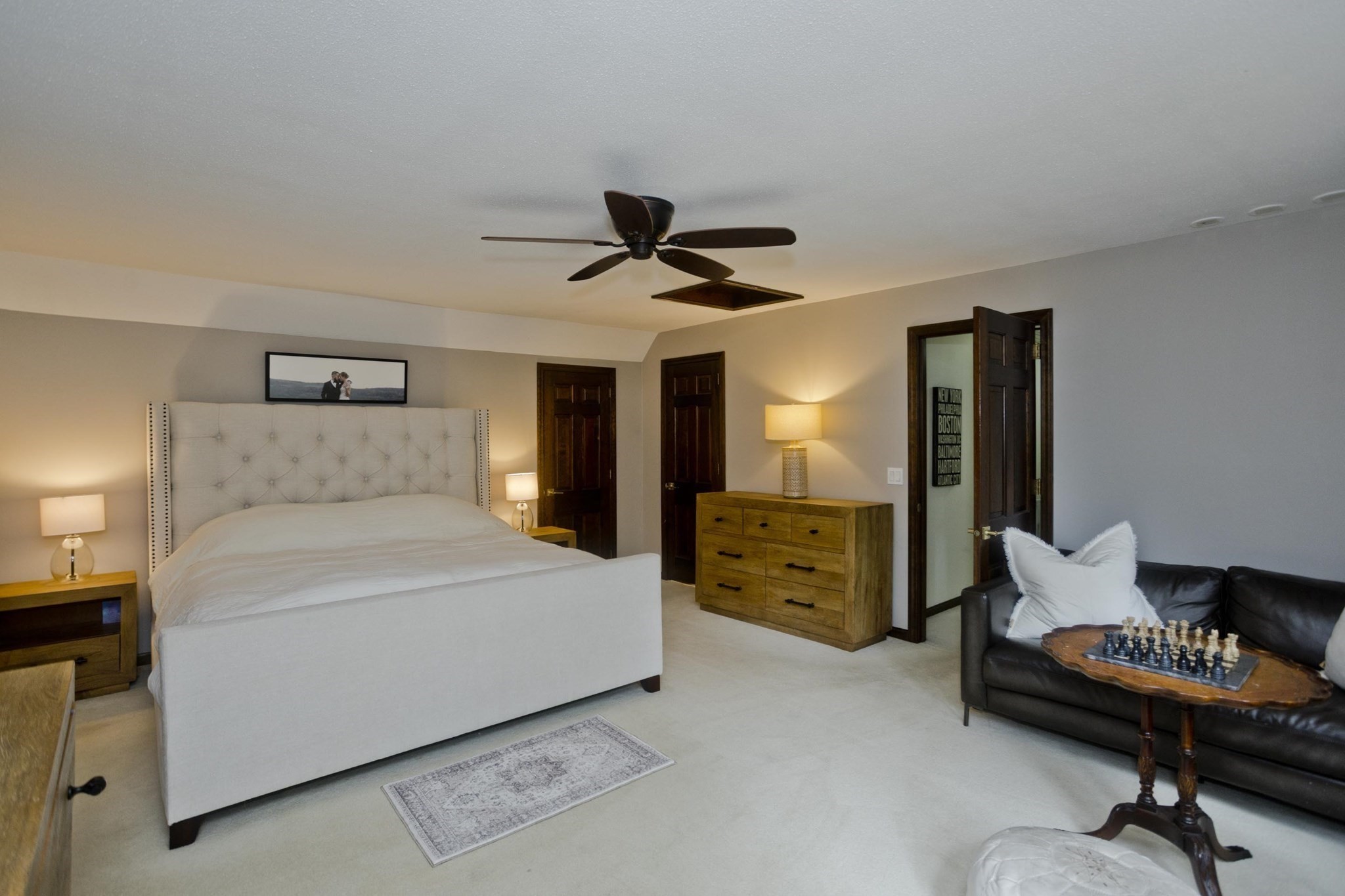 Bedroom with a large white upholstered bed, wood dresser, ceiling fan, and sofa with a chessboard.
