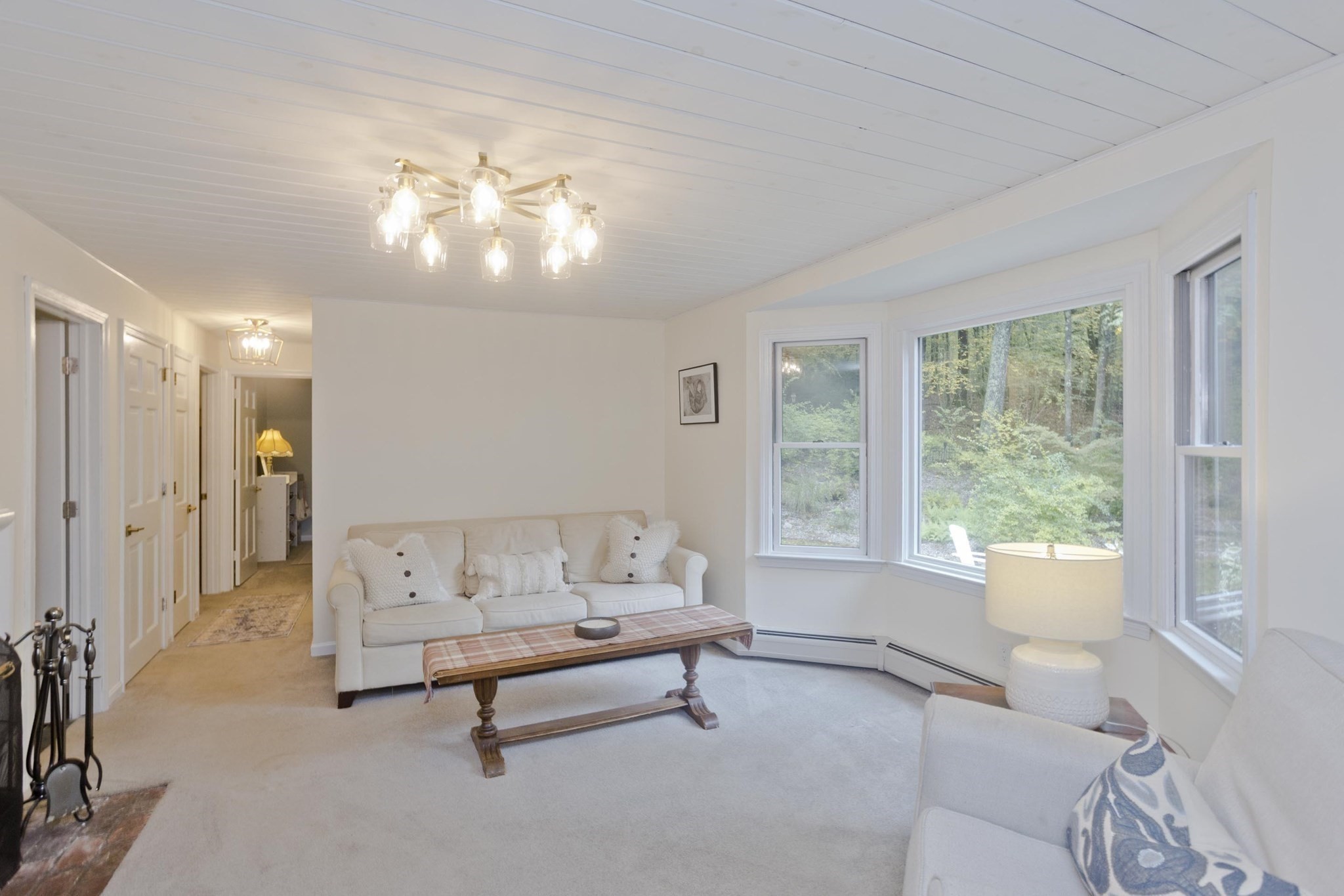 Living room with white sofas and a plaid coffee table beside large windows overlooking trees.