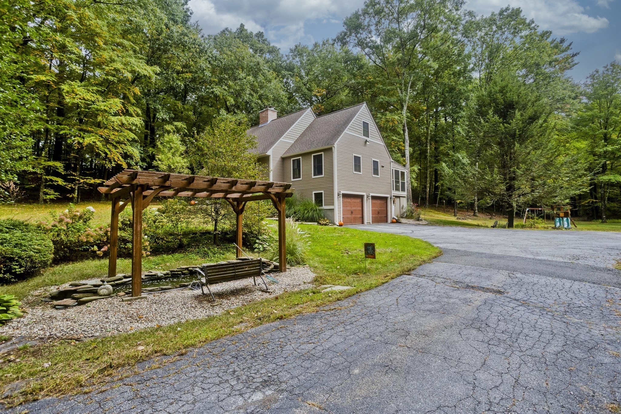 Beige house with two garage doors, beside a pergola-covered pond and bench on a wooded lot.