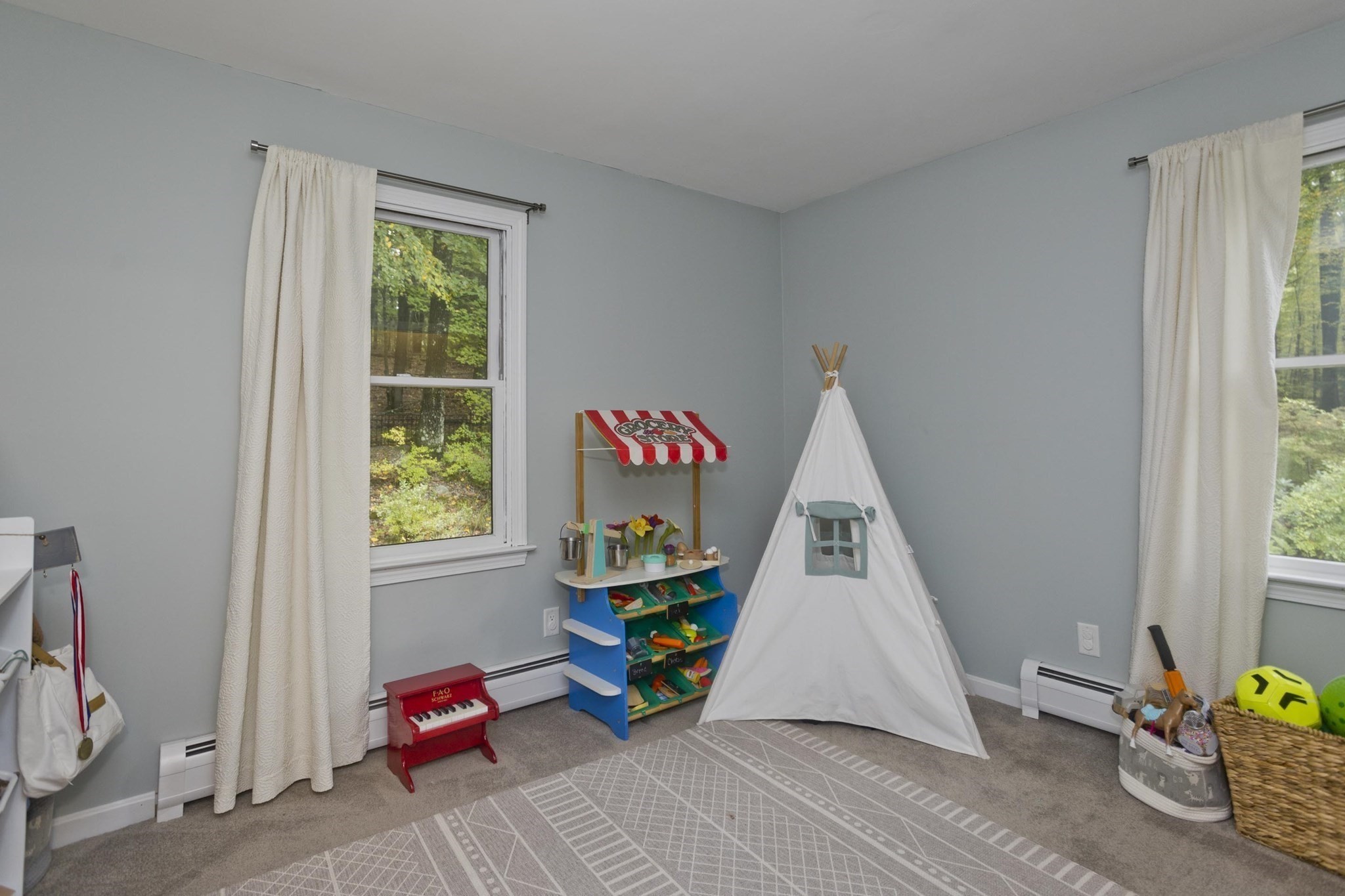 Children’s playroom with a white teepee, toy market stand, and red piano between two windows.
