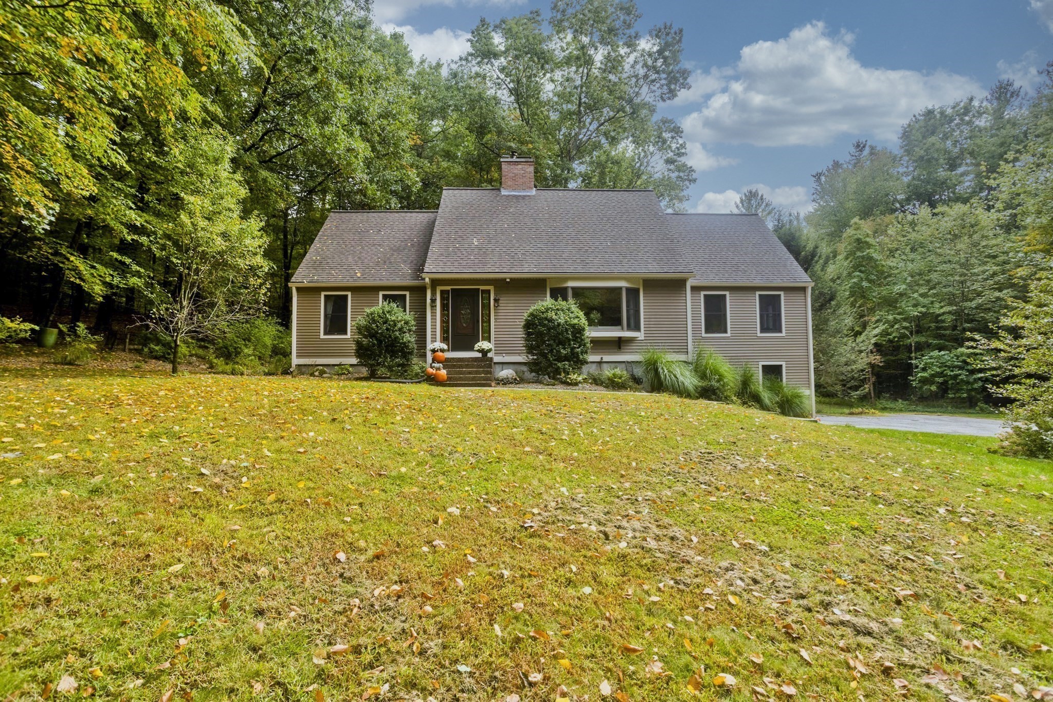 Beige house with a brick chimney, front steps decorated with pumpkins, and a leaf-strewn lawn.