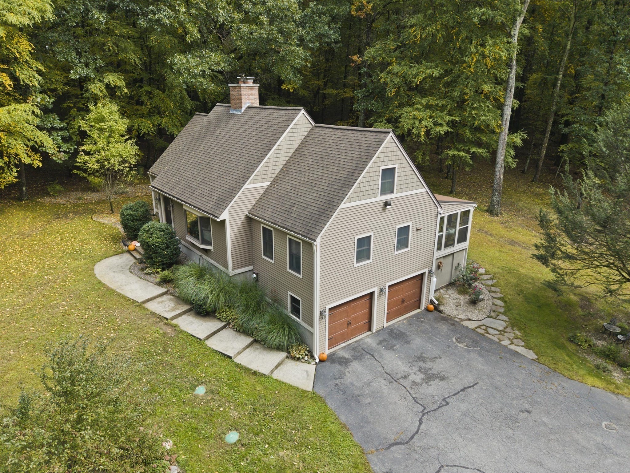 Aerial view of a beige gabled house with two garage doors, a curved walkway, and wooded autumn grounds.