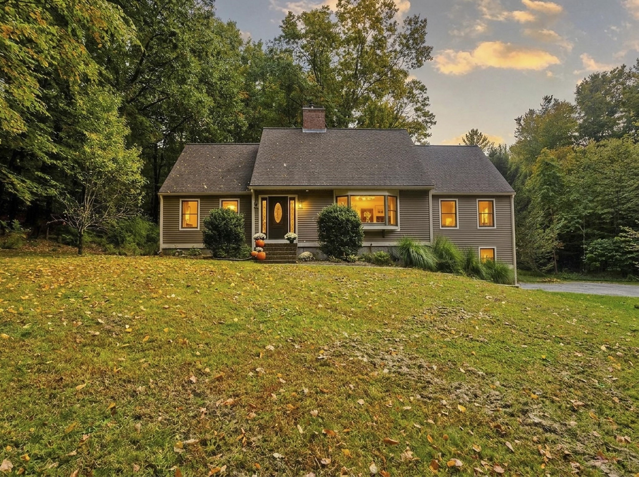 Gray-sided home with warmly lit windows, a leaf-covered lawn, and trees at dusk.