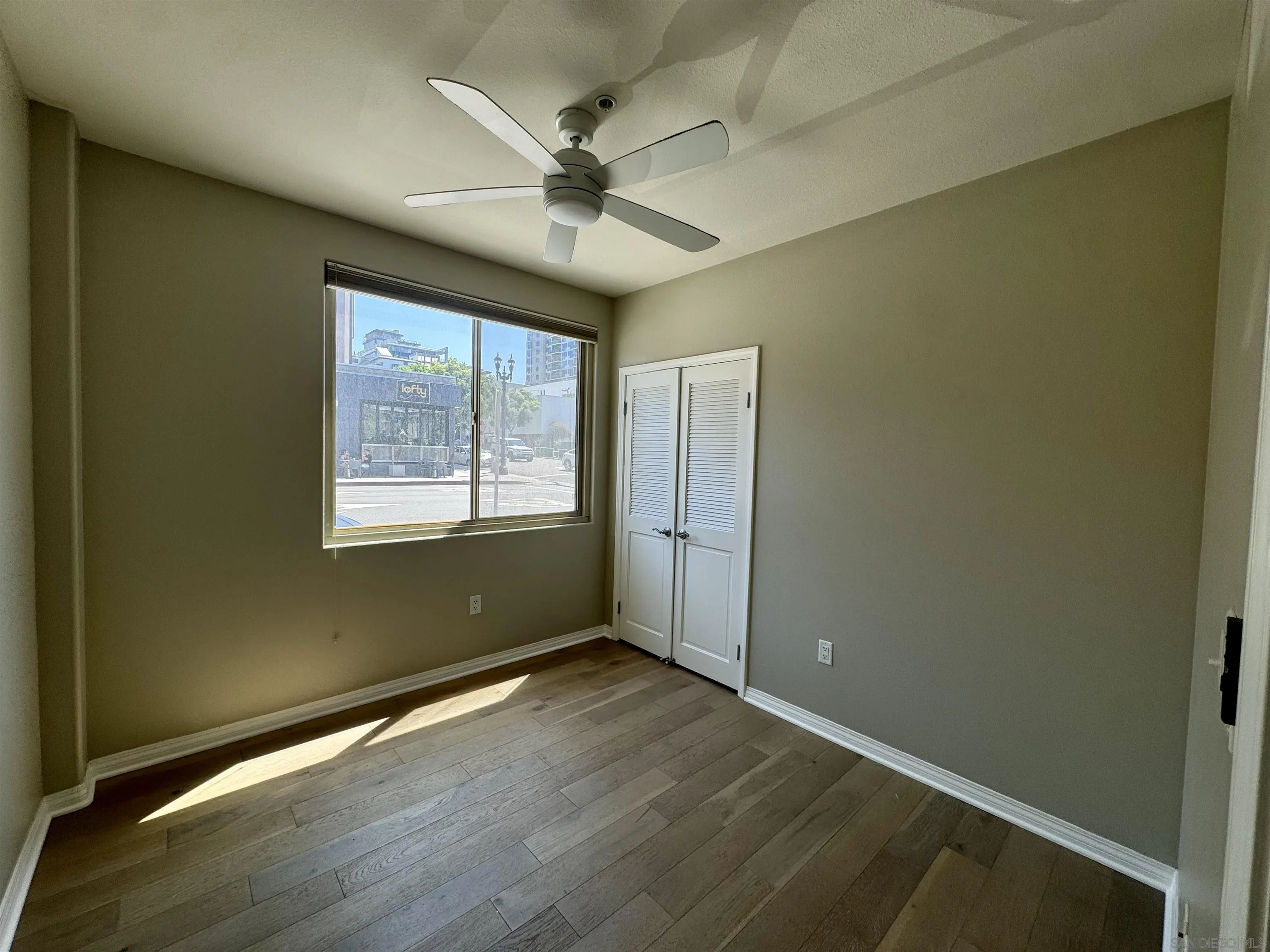 Empty room with wood flooring, ceiling fan, city-view window, and louvered closet doors.