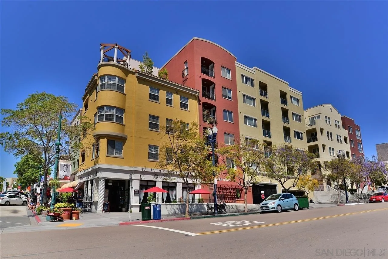 Colorful multistory condominium building with balconies and storefronts on a sunny urban corner.