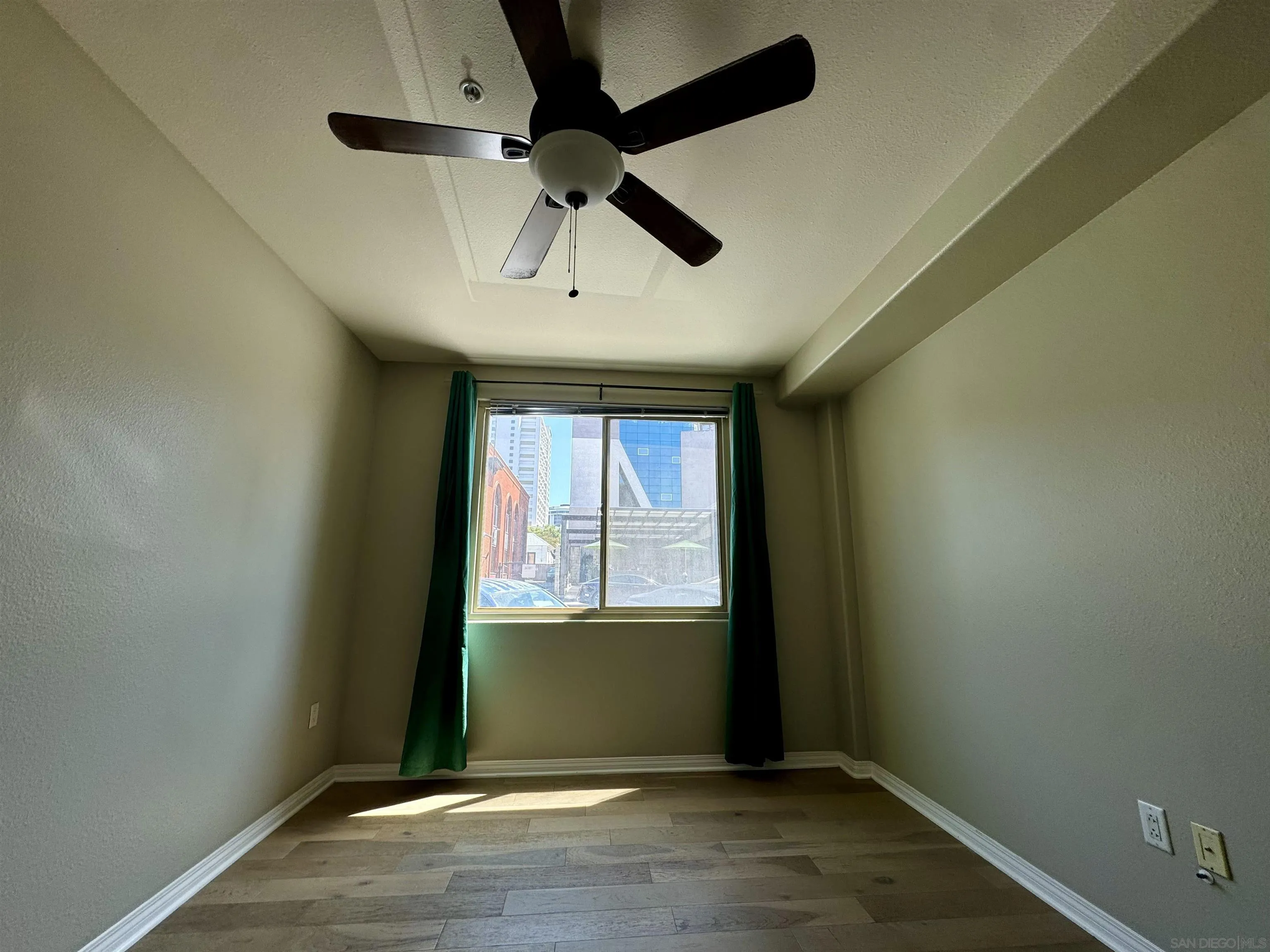 Empty bedroom with wood-look flooring, a ceiling fan, and green curtains around a city-facing window.