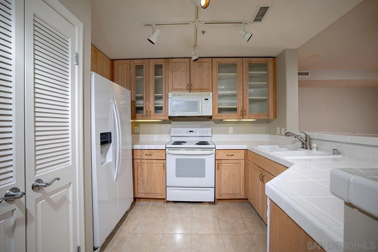 Kitchen with light wood cabinets, white appliances, tiled counters, and a double sink.