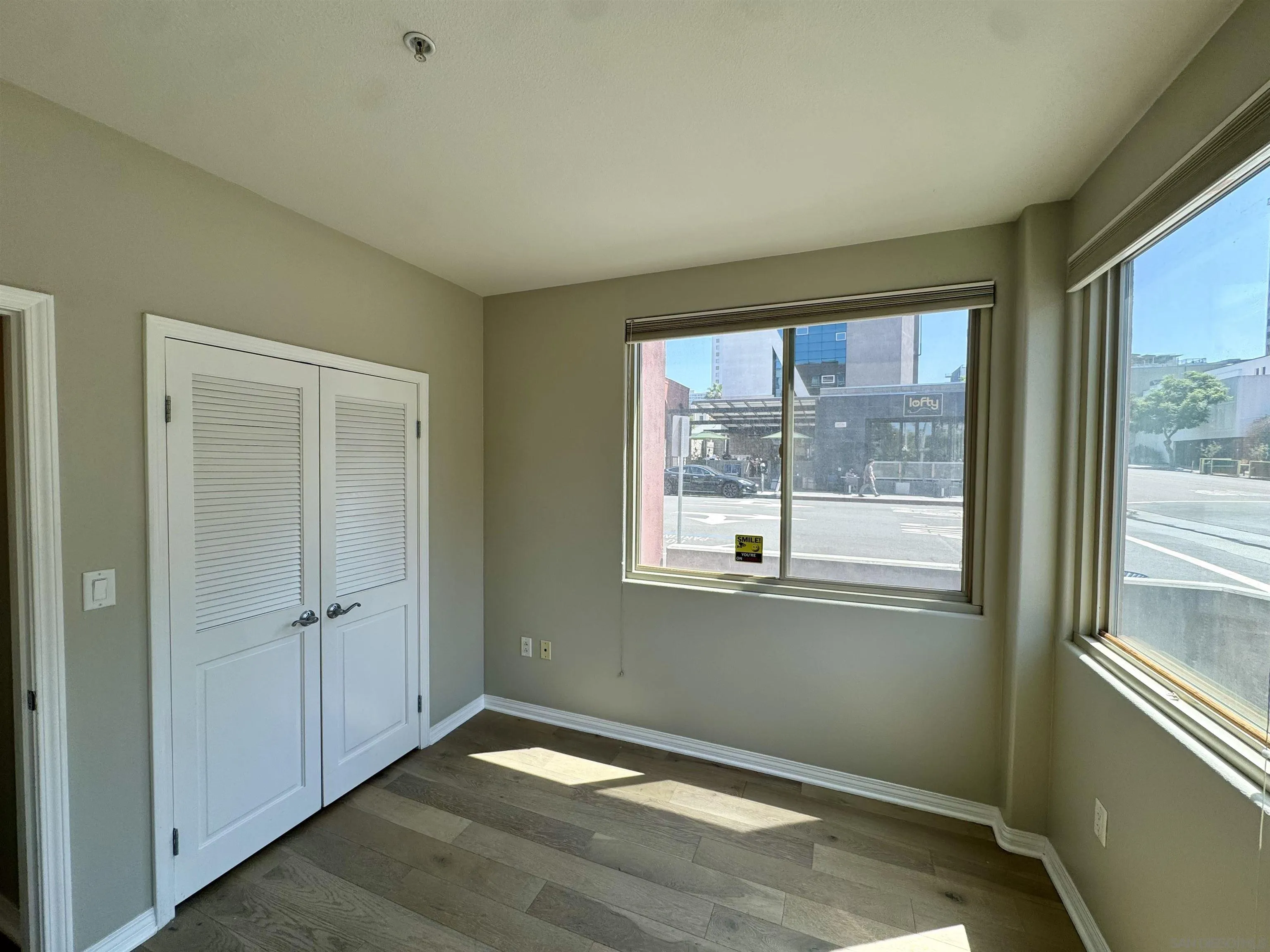 Empty room with wood flooring, louvered doors, and large windows overlooking the street.