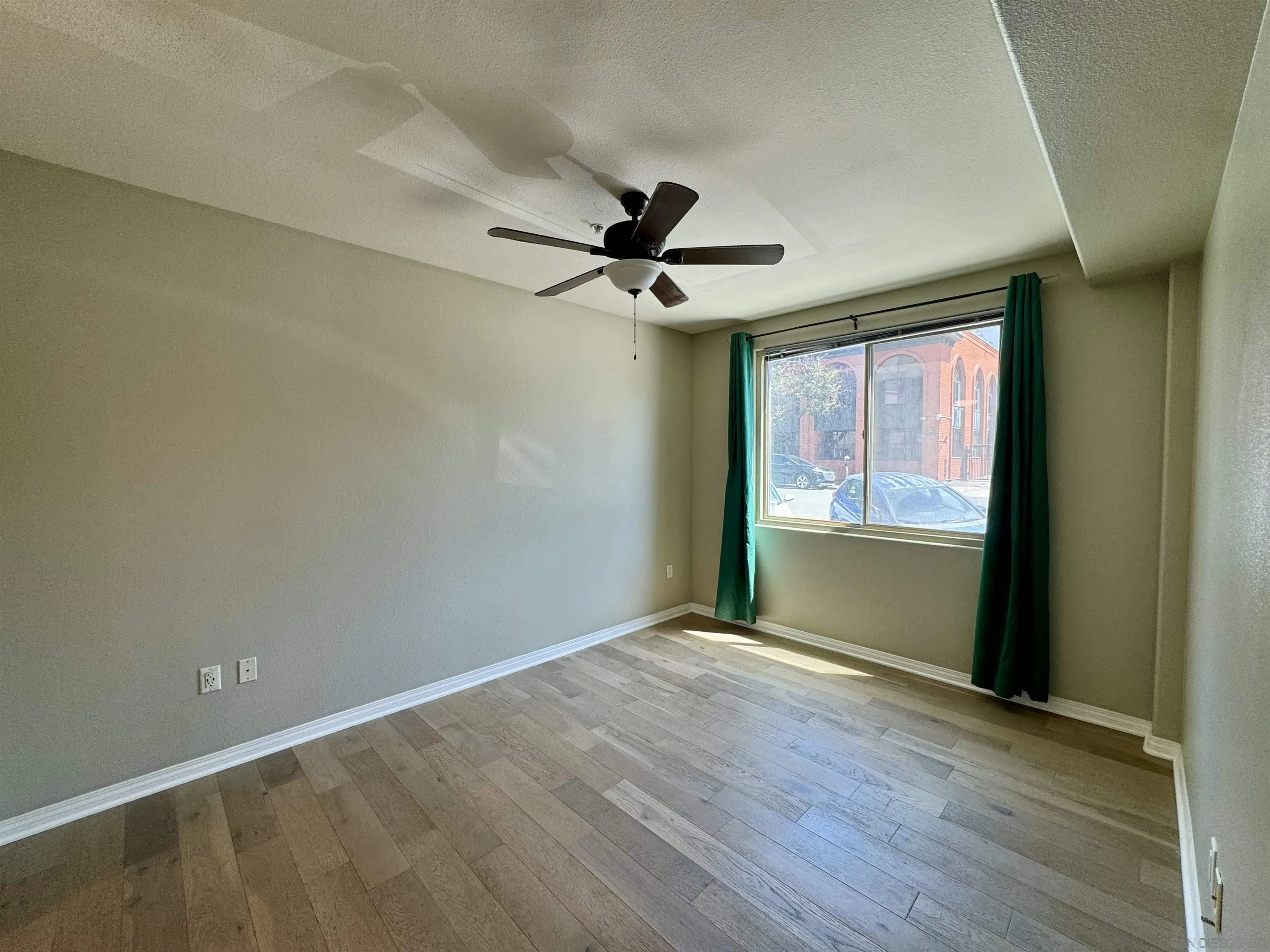 Empty bedroom with wood-look flooring, a ceiling fan, and green curtains framing a sunny window.