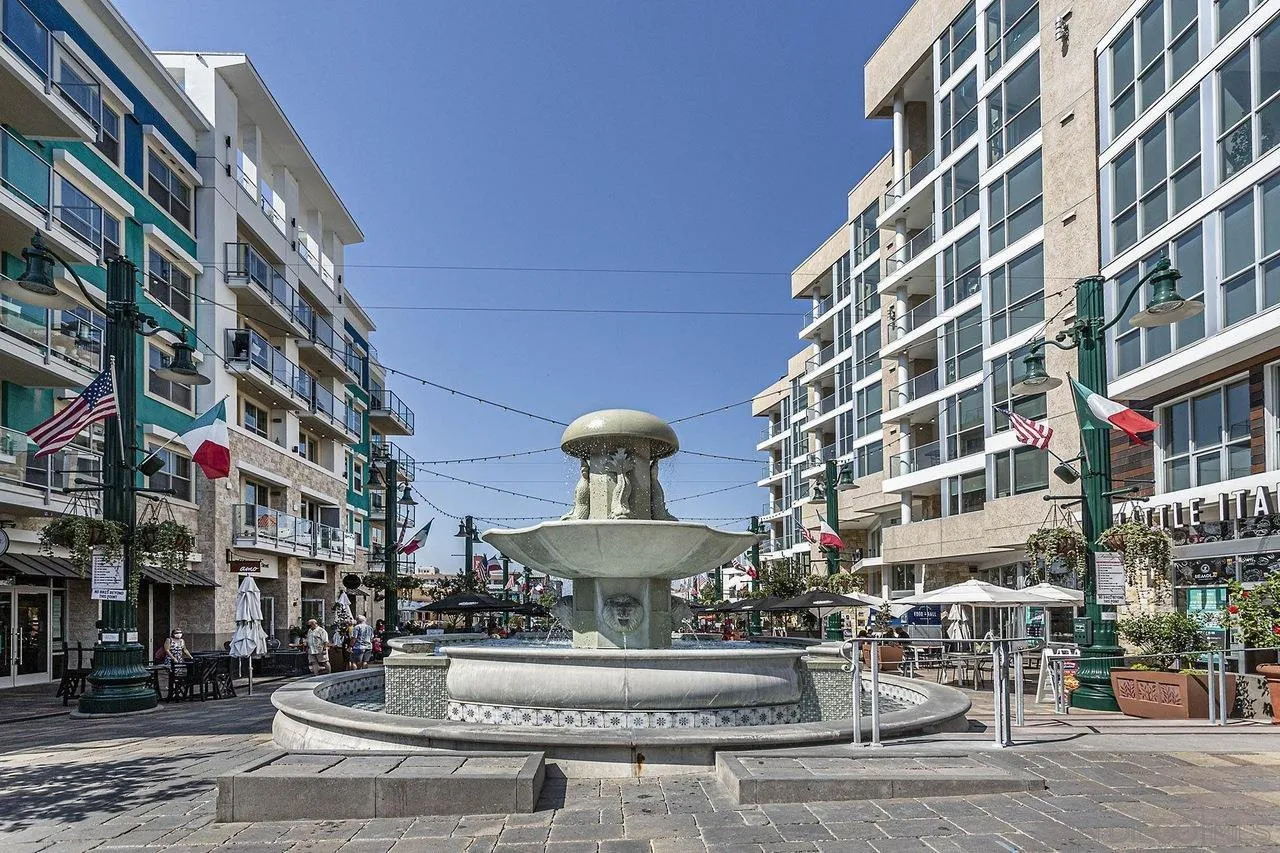 Pedestrian plaza with a stone fountain, outdoor dining, and modern buildings in Little Italy.