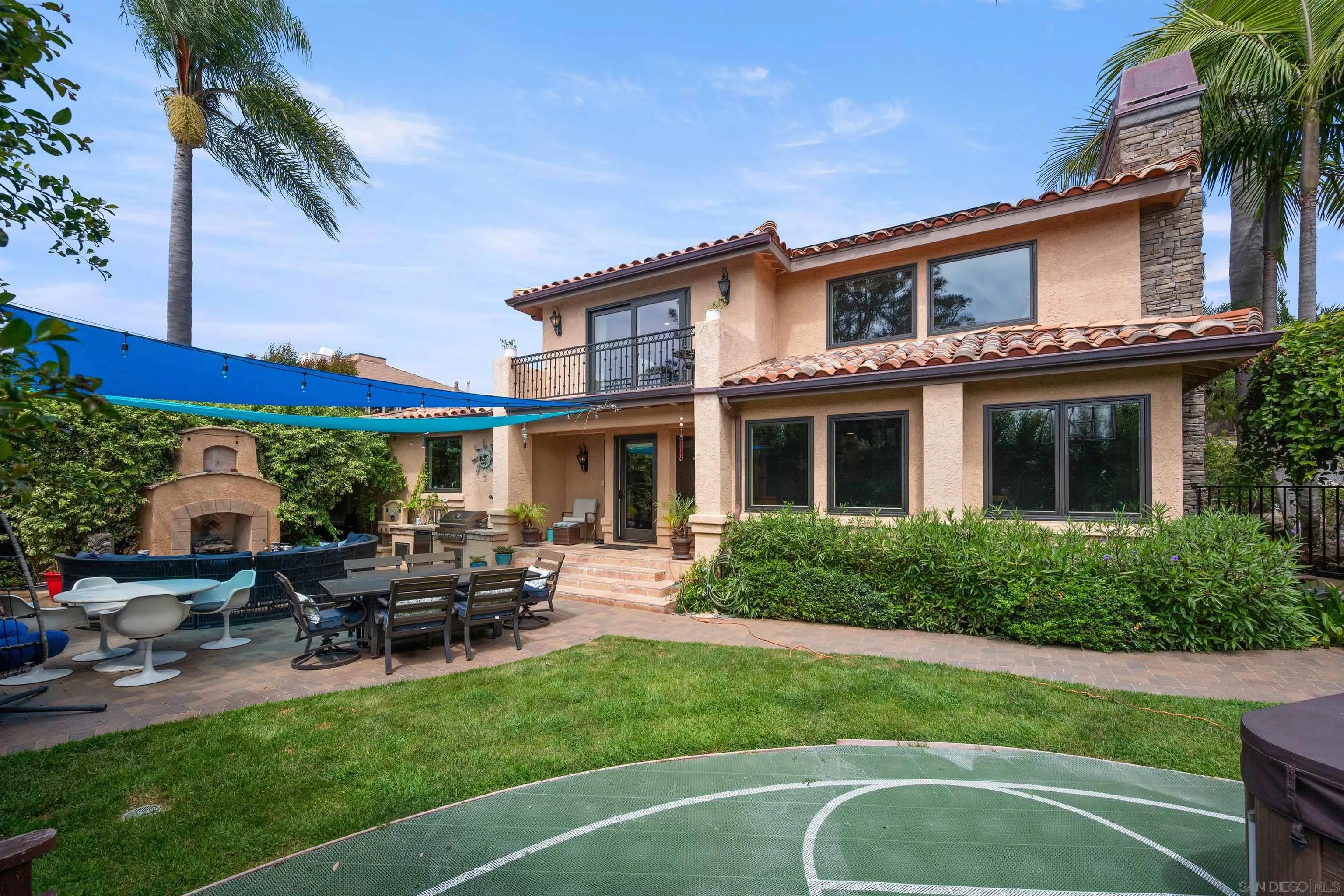 Home’s backyard with a tiled patio, outdoor fireplace, shade sails, lawn, and small sport court.
