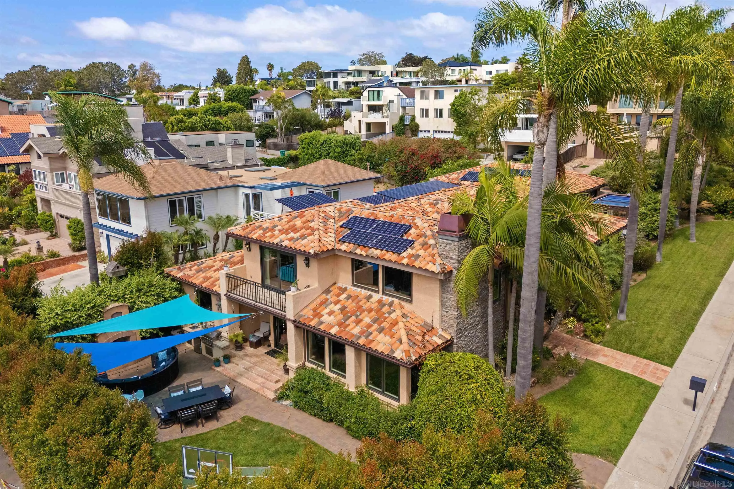 Aerial view of a Del Mar home with solar panels, palm trees, and a pool beneath blue shade sails.