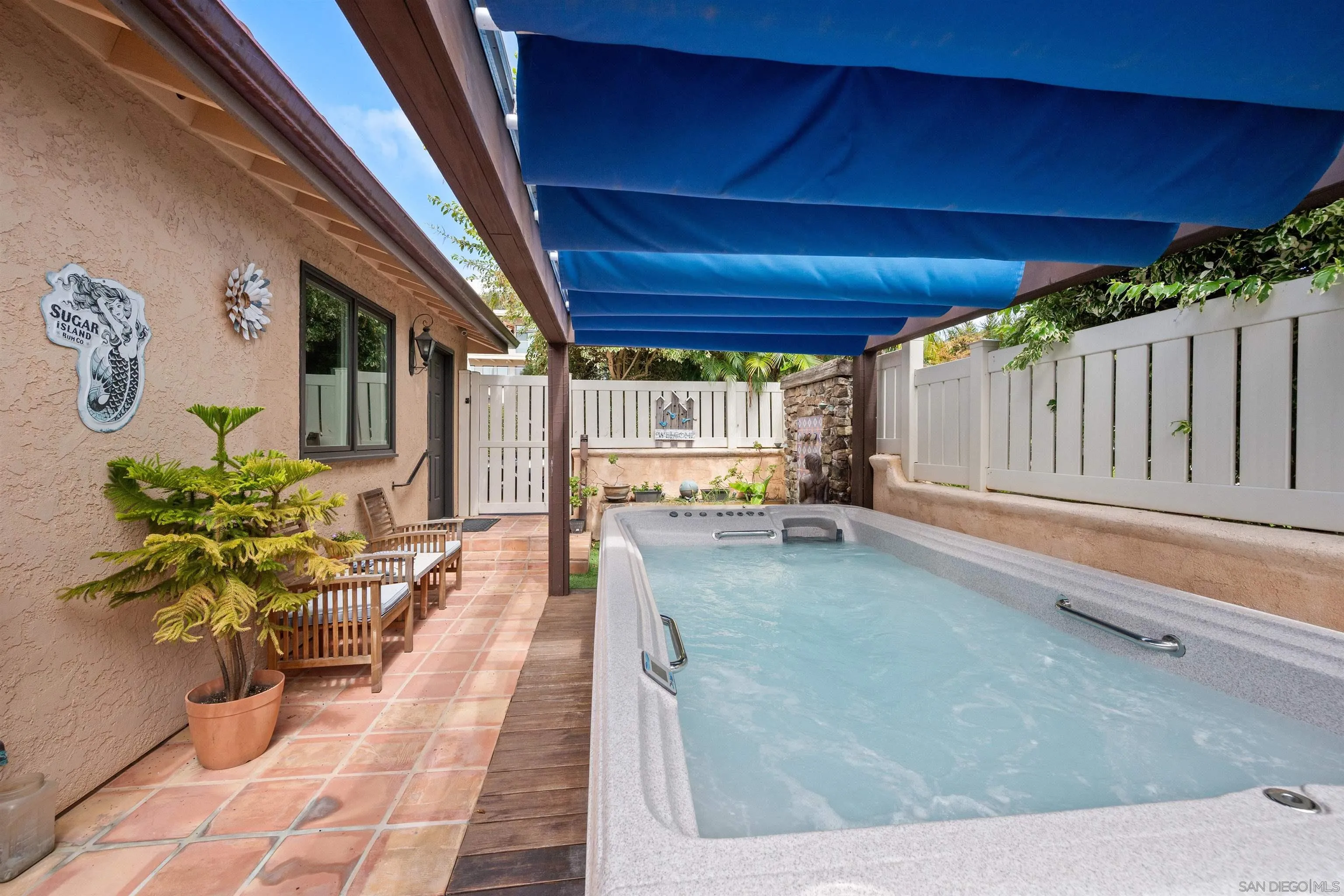 Outdoor spa beneath a blue retractable awning, beside a tiled patio with wooden seating and potted plants.