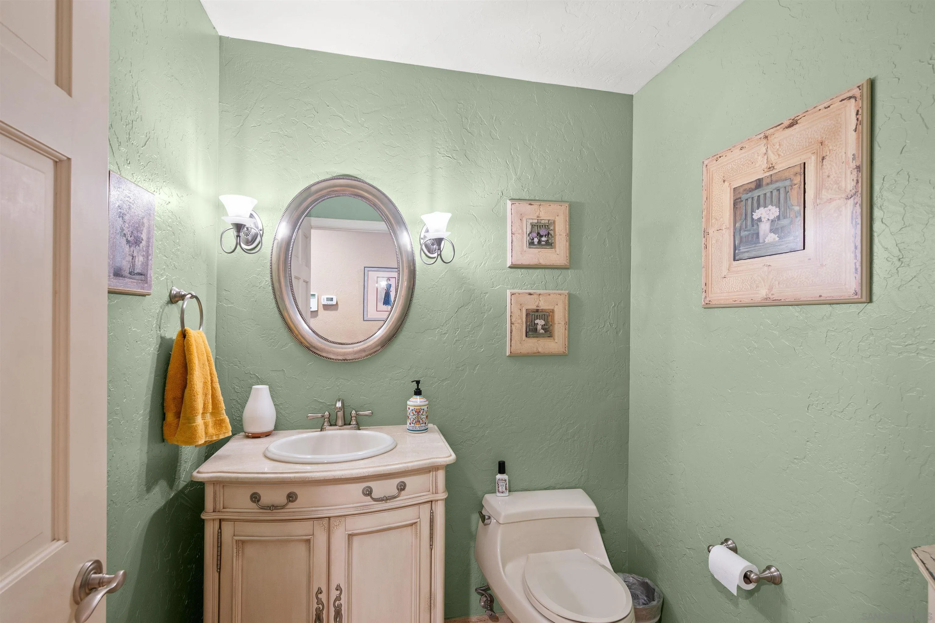 Bathroom with sage-green walls, a cream vanity, oval mirror, and framed artwork.