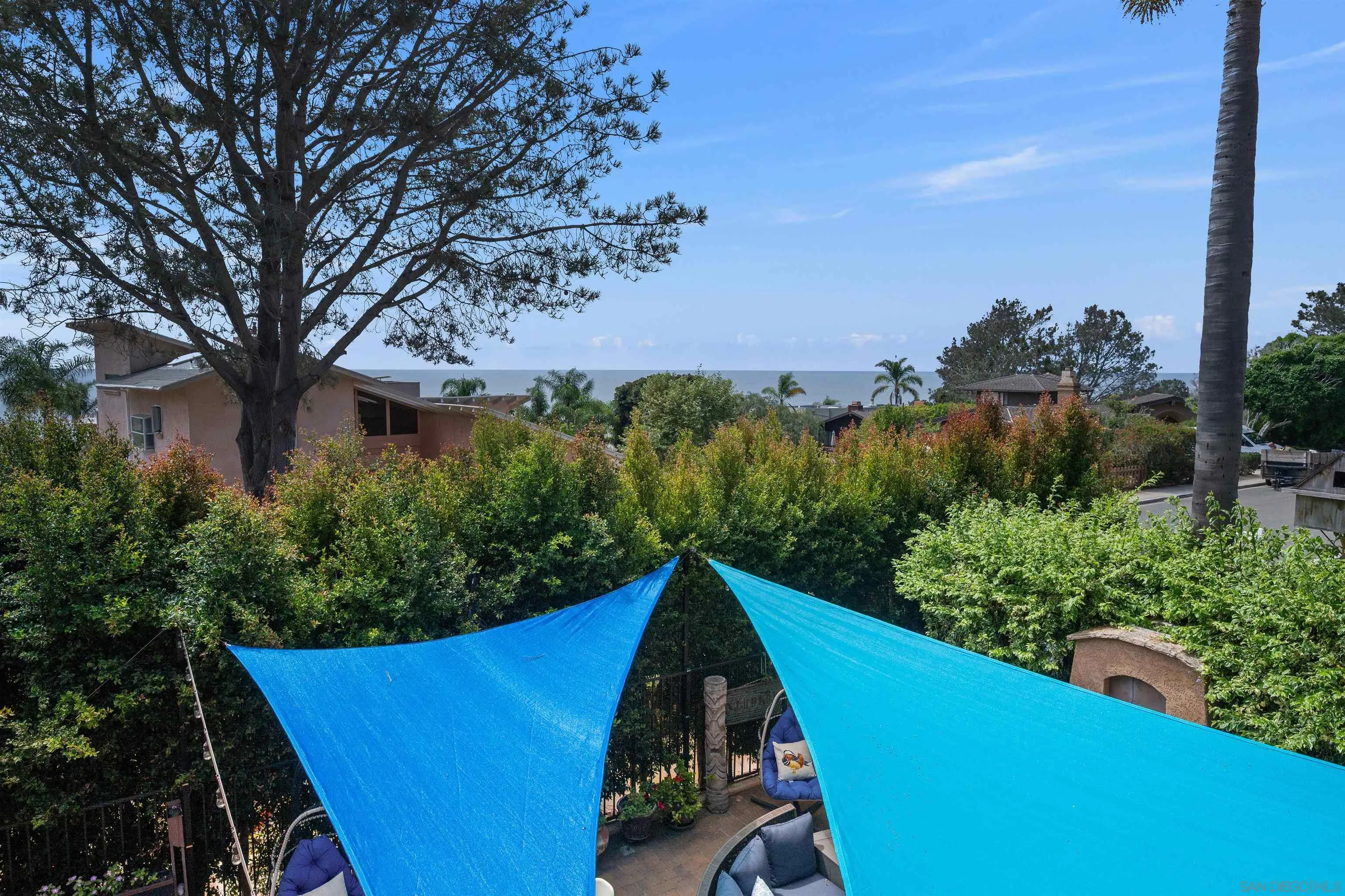 Ocean-view patio with blue shade sails, leafy hedges, and outdoor seating.