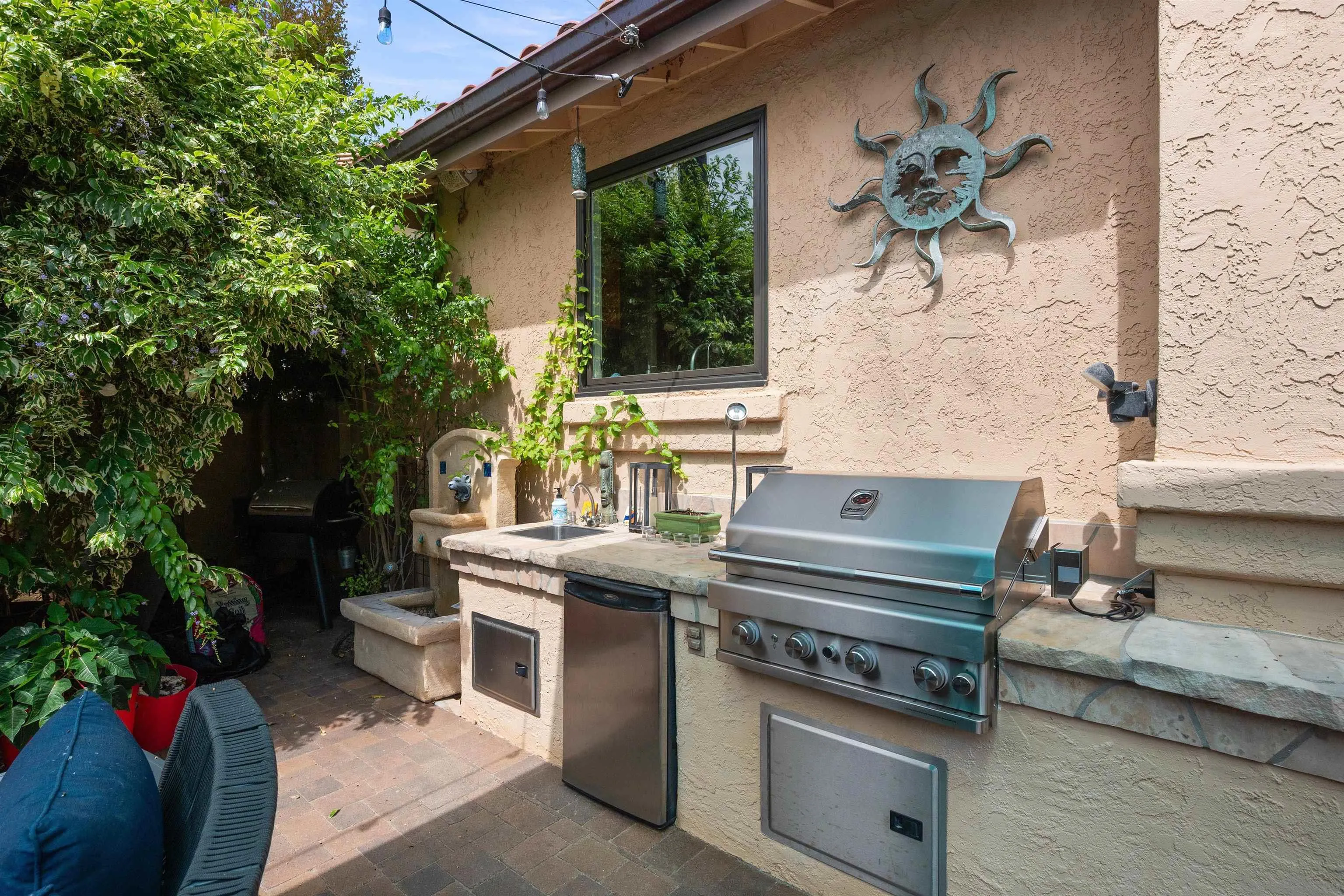 Outdoor kitchen with a stainless-steel grill, sink, and refrigerator beside lush greenery.