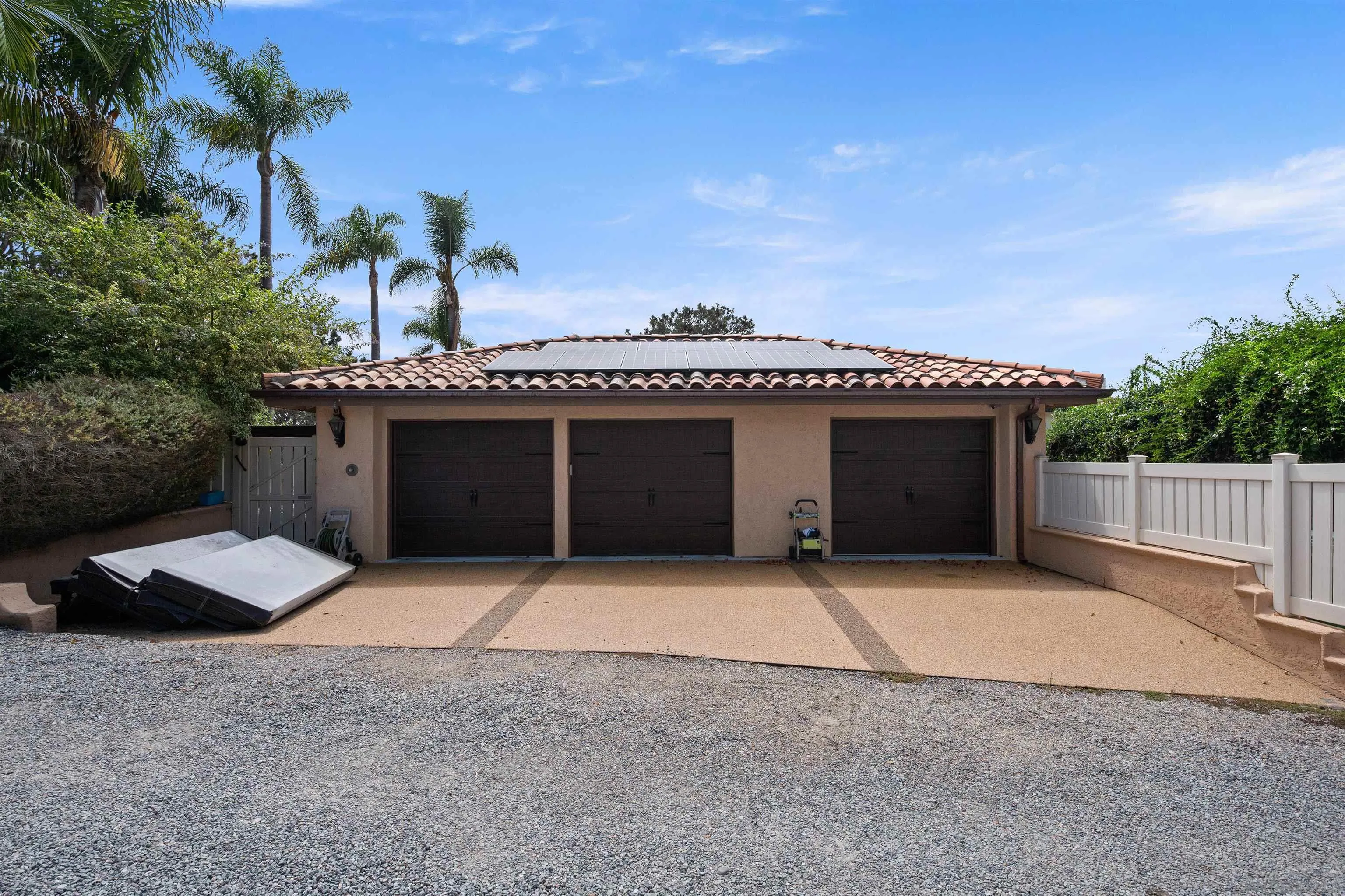 Three-car garage with rooftop solar panels, a gravel driveway, white fencing, and palm trees.