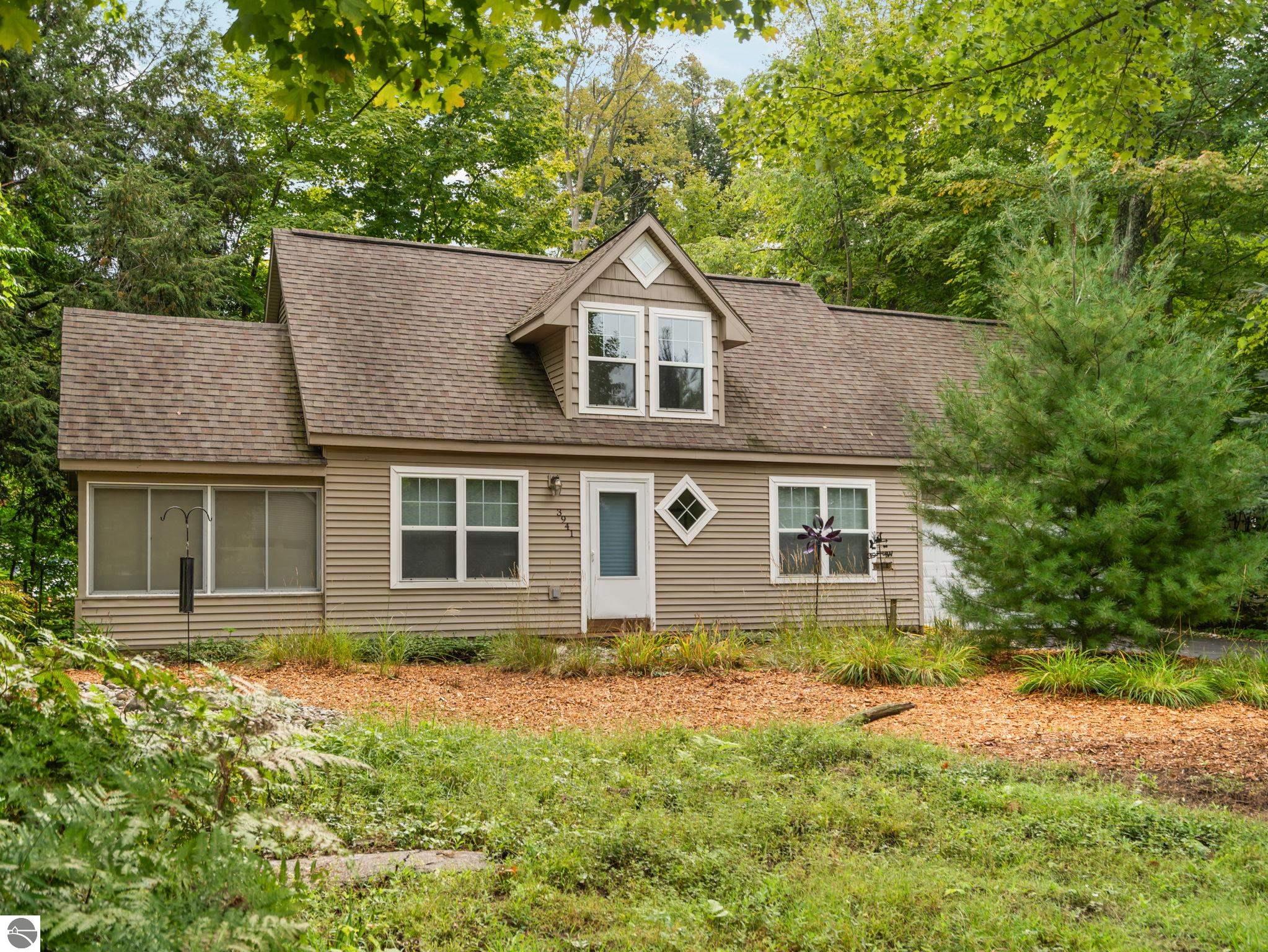 Beige two-story house with a prominent roof dormer, surrounded by mature trees and landscaping.