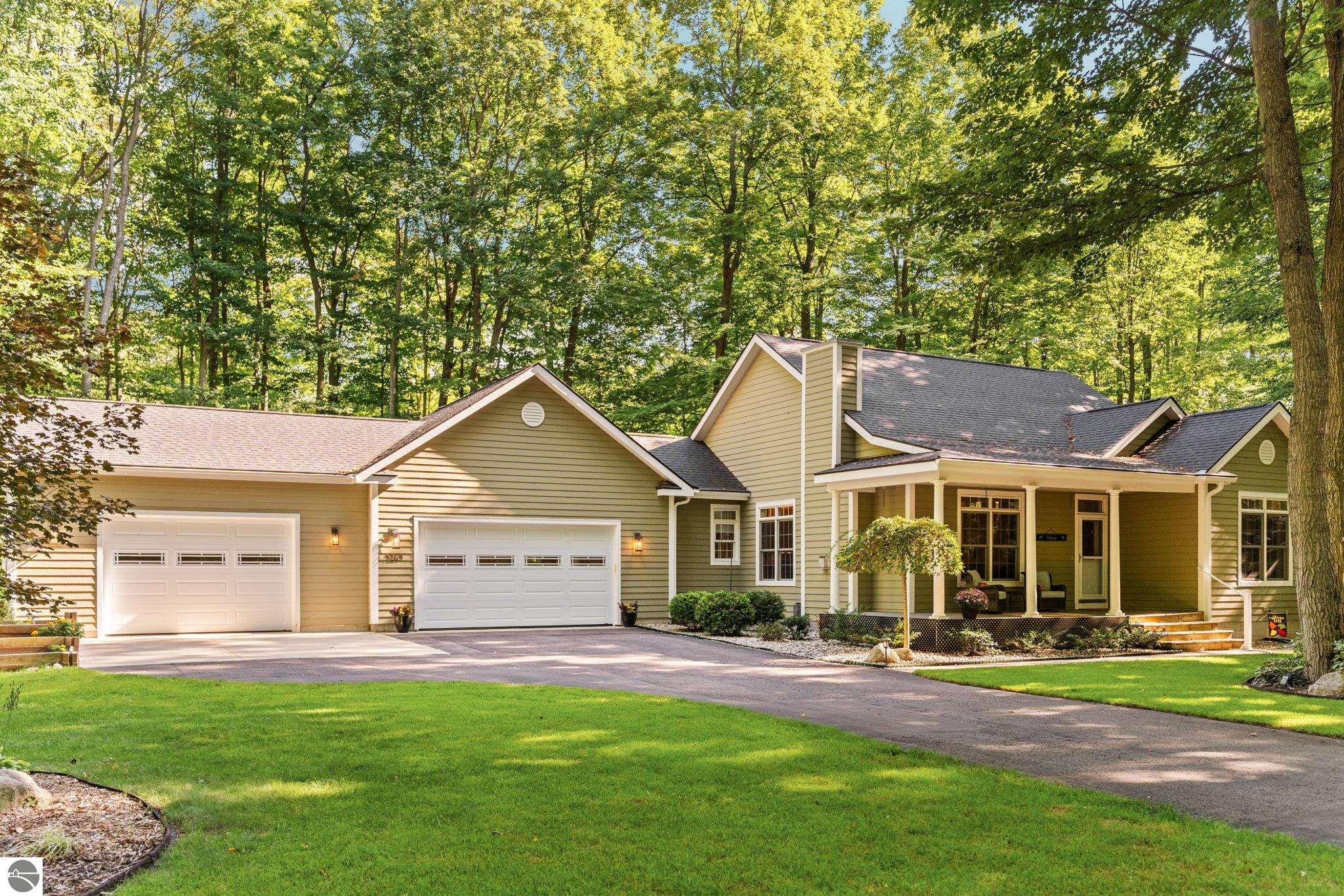 Beige home with a covered porch and two garage doors, surrounded by mature trees.