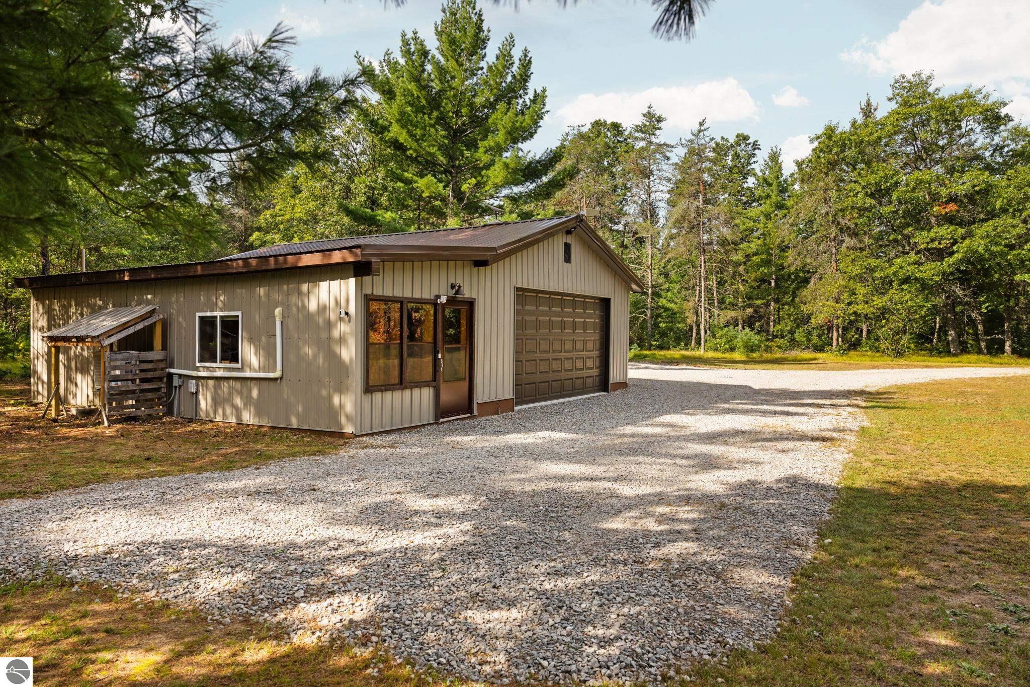 Beige metal-sided outbuilding with a garage door beside a broad gravel driveway and mature trees.
