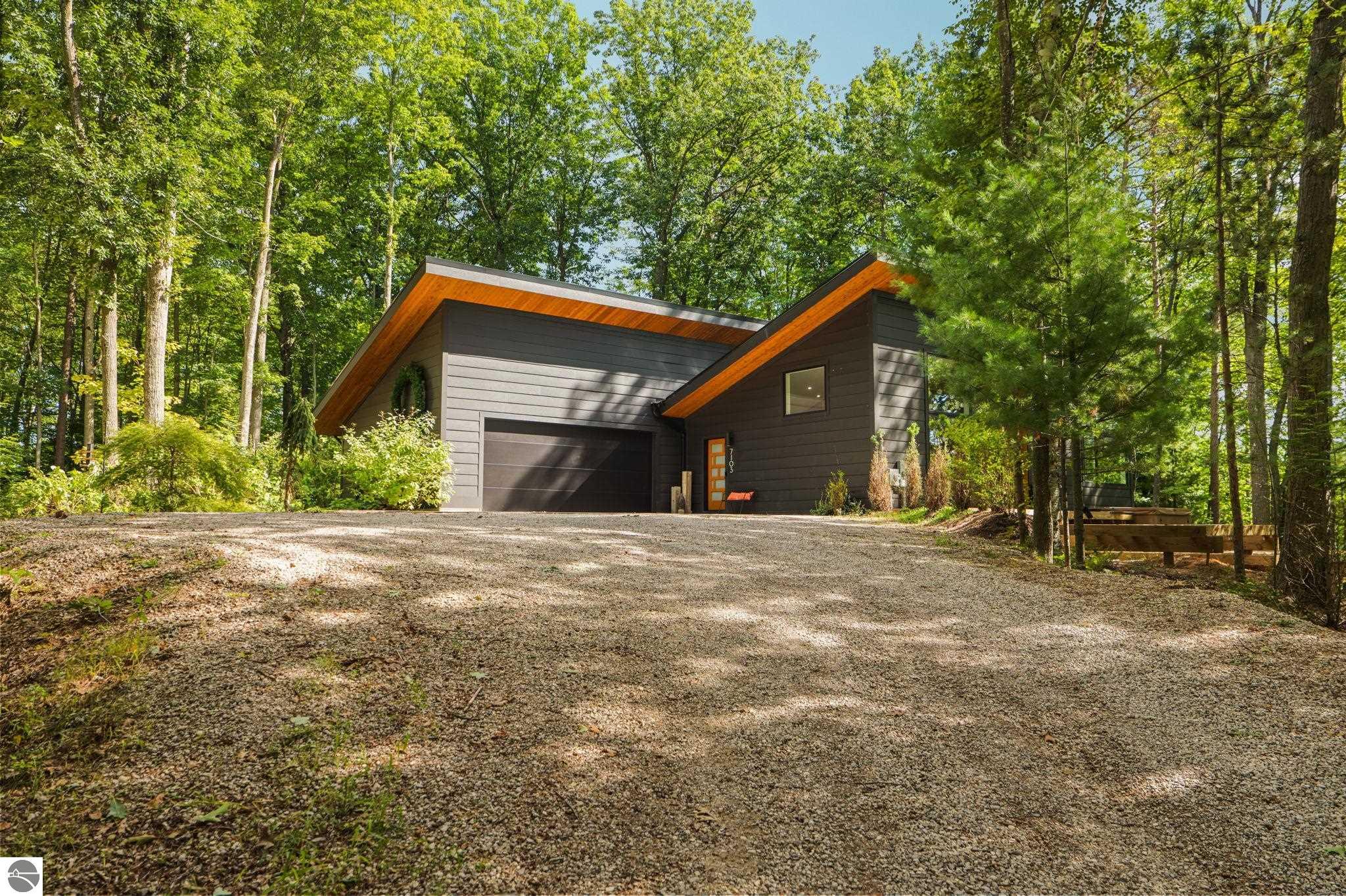 Modern charcoal home with warm wood roof soffits, a gravel drive, and a wooded setting.