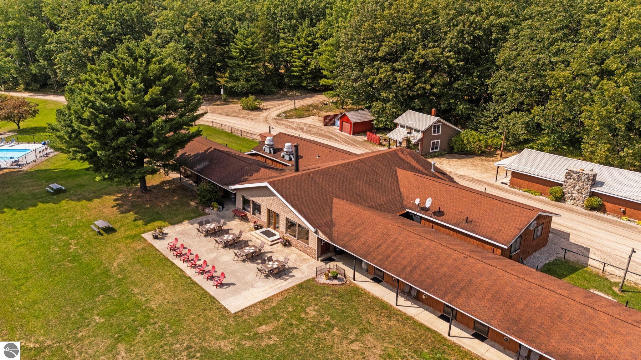 Aerial view of a wooded property with a large building, patio seating, lawn, and swimming pool.
