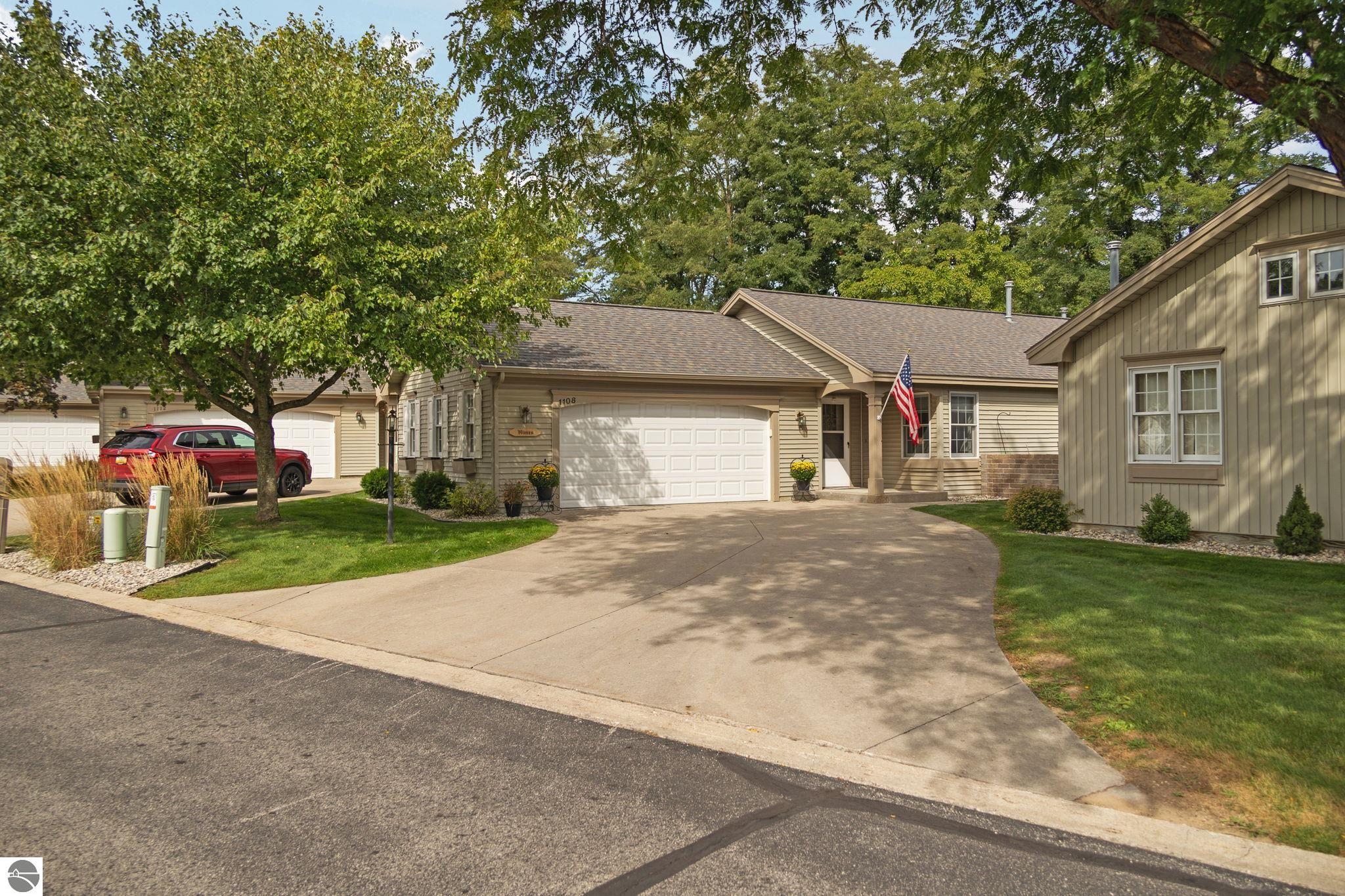 Tan attached home with a double garage, curved driveway, mature trees, and an American flag.