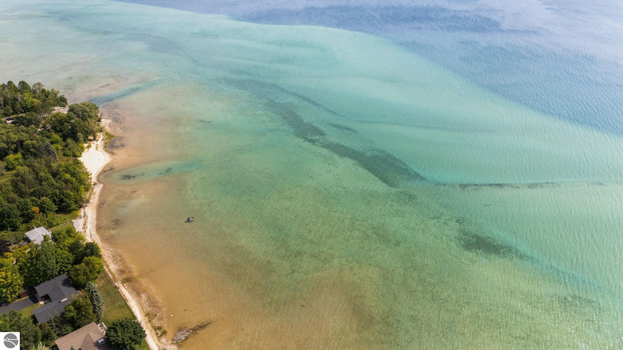Aerial view of a wooded shoreline and homes beside clear, shallow turquoise water.