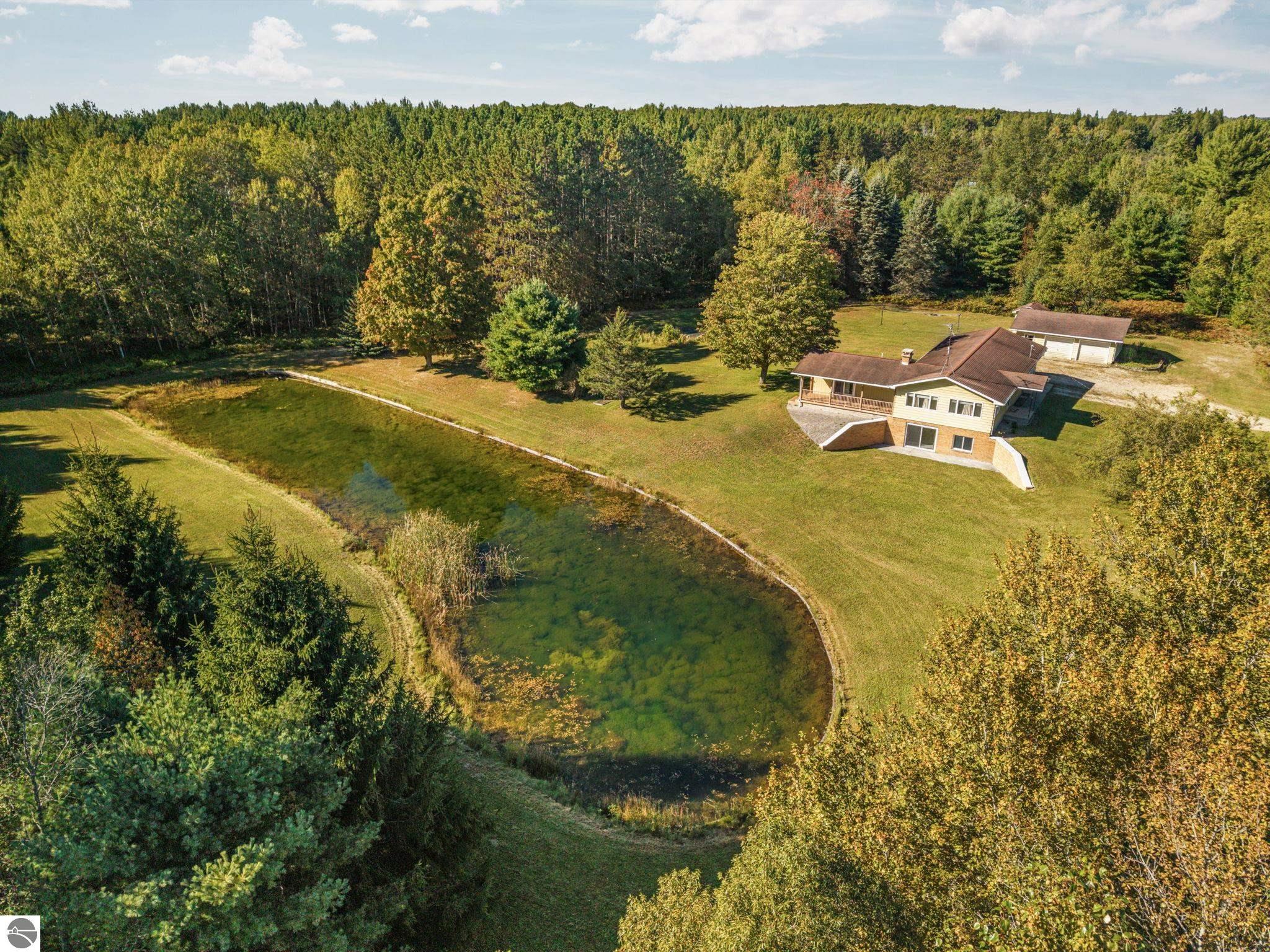 Aerial view of a home beside a pond, with a detached garage and wooded acreage.