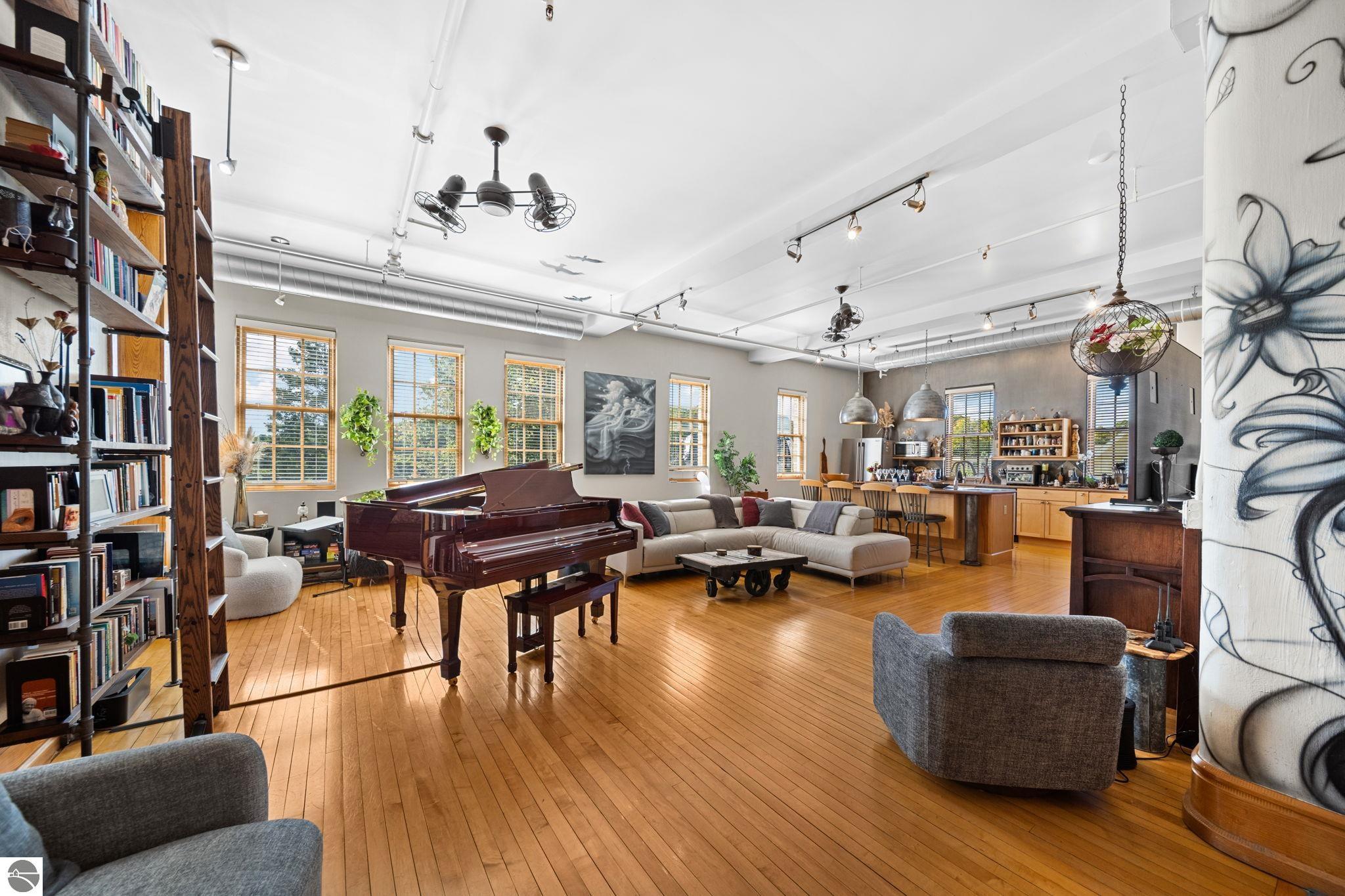 Open-plan living room with a grand piano, bookshelves, wood floors, and an adjoining kitchen.