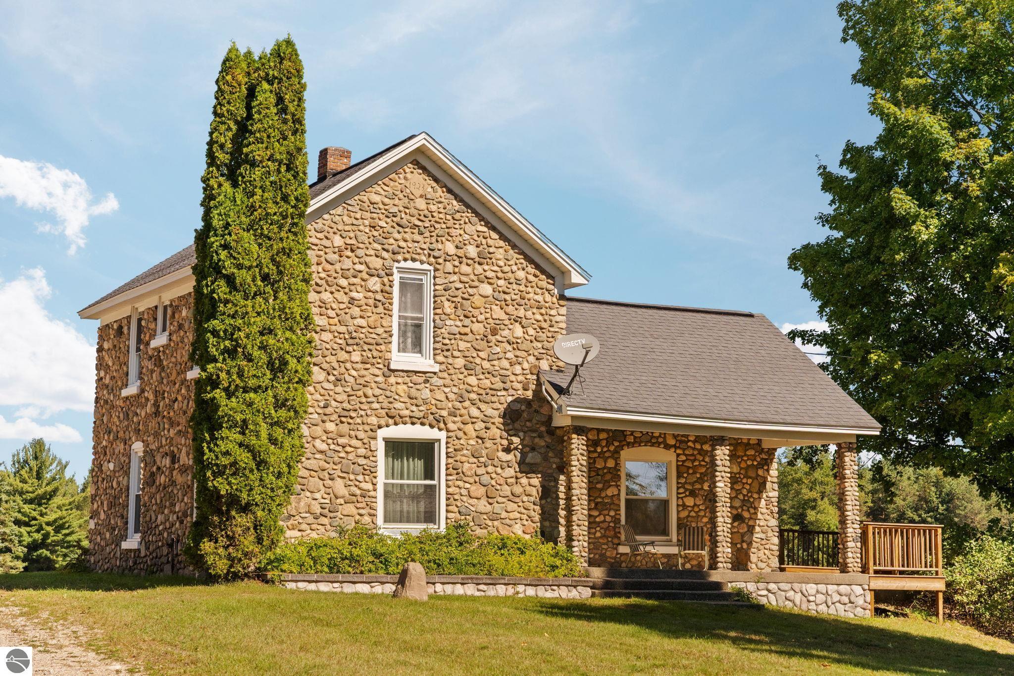 Two-story stone house with a covered porch, attached deck, and tall evergreen in front.
