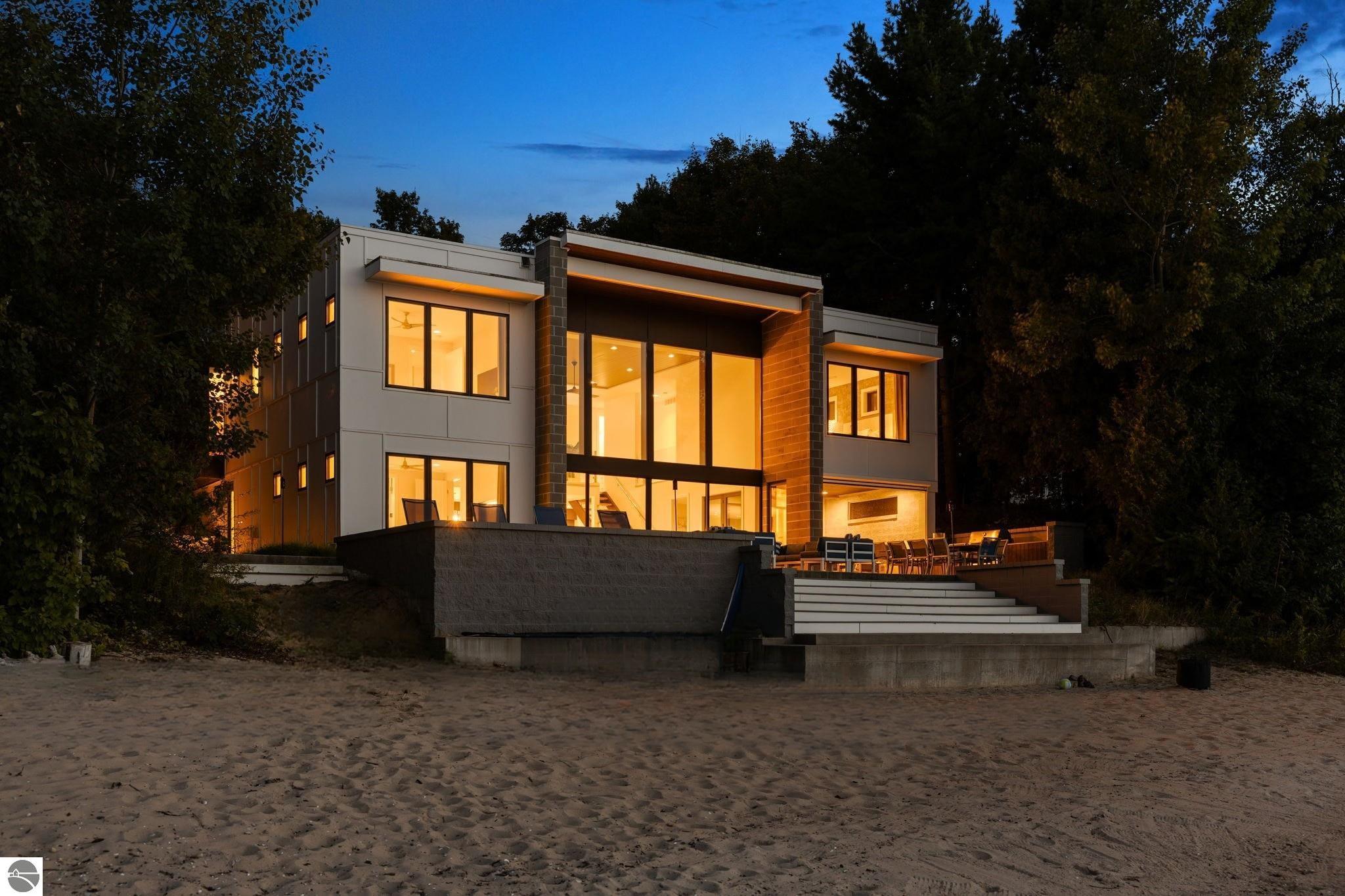 Modern multi-story beachside home glowing at dusk, with a broad patio and steps leading to the sand.
