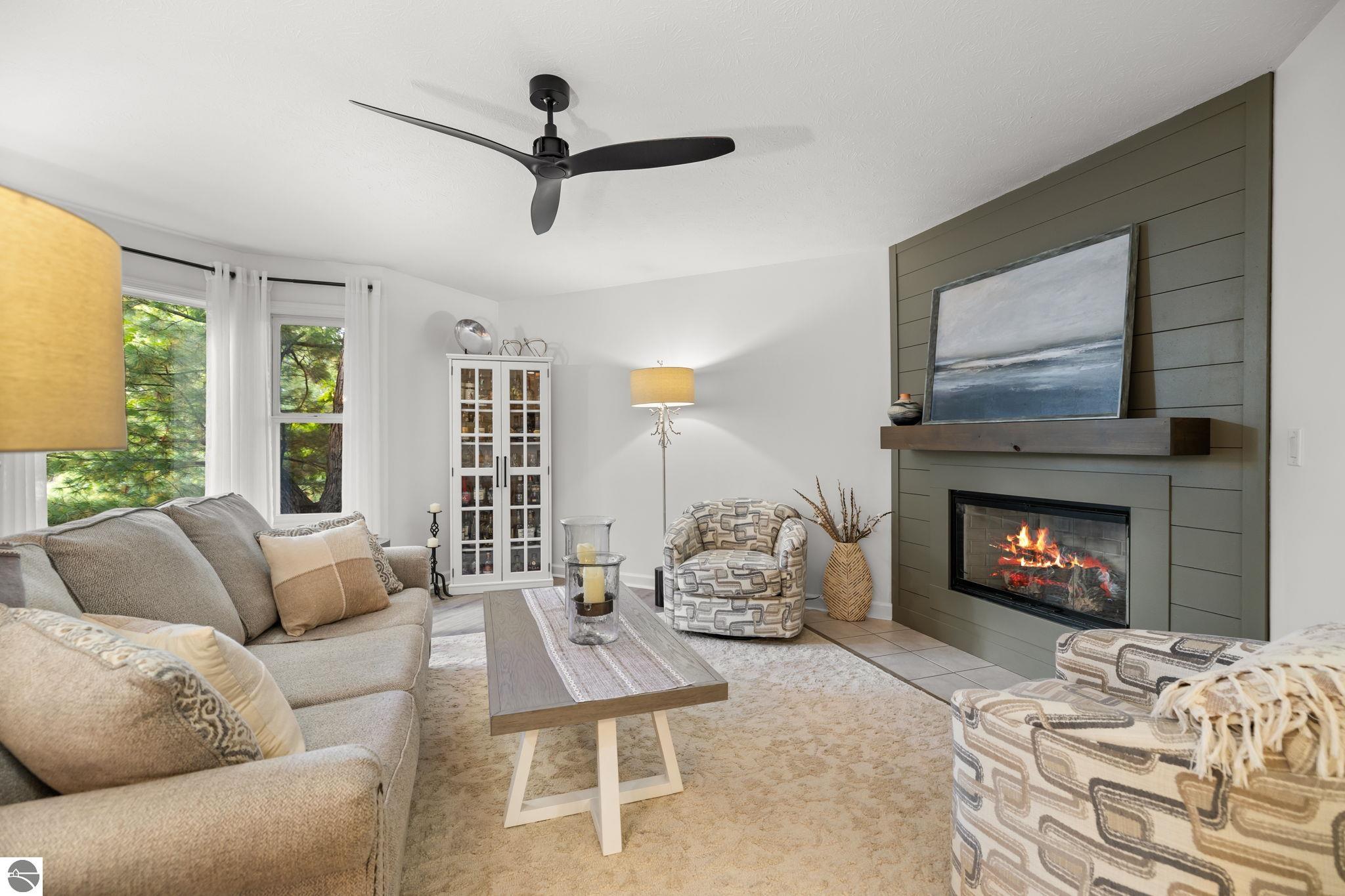 Staged living room with a lit fireplace, gray sofa, patterned chairs, and windows overlooking trees.