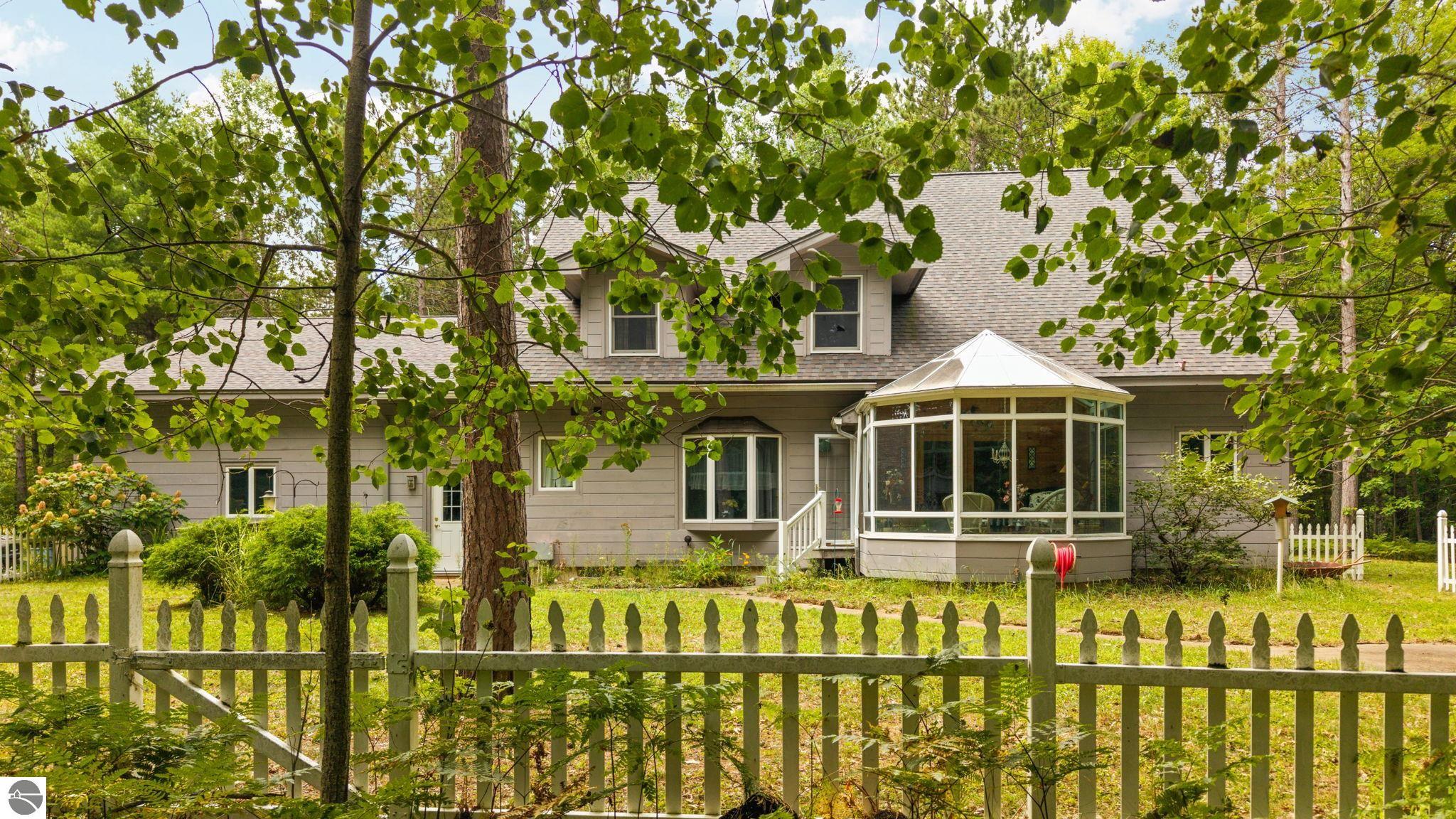 Gray two-story house with a glass-enclosed sunroom behind a white picket fence.