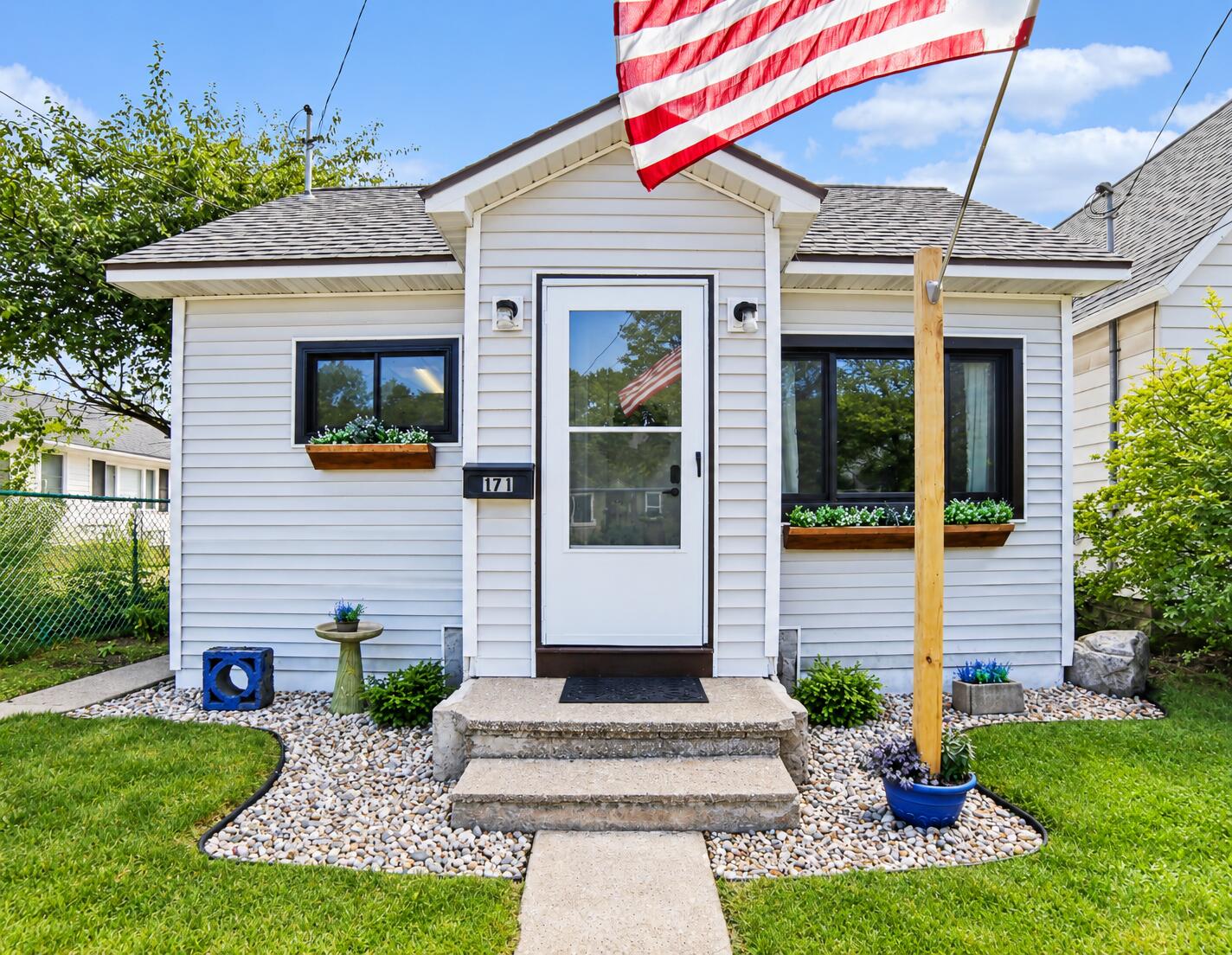White-sided house entrance with stone steps, flower boxes, black-framed windows, and an American flag.