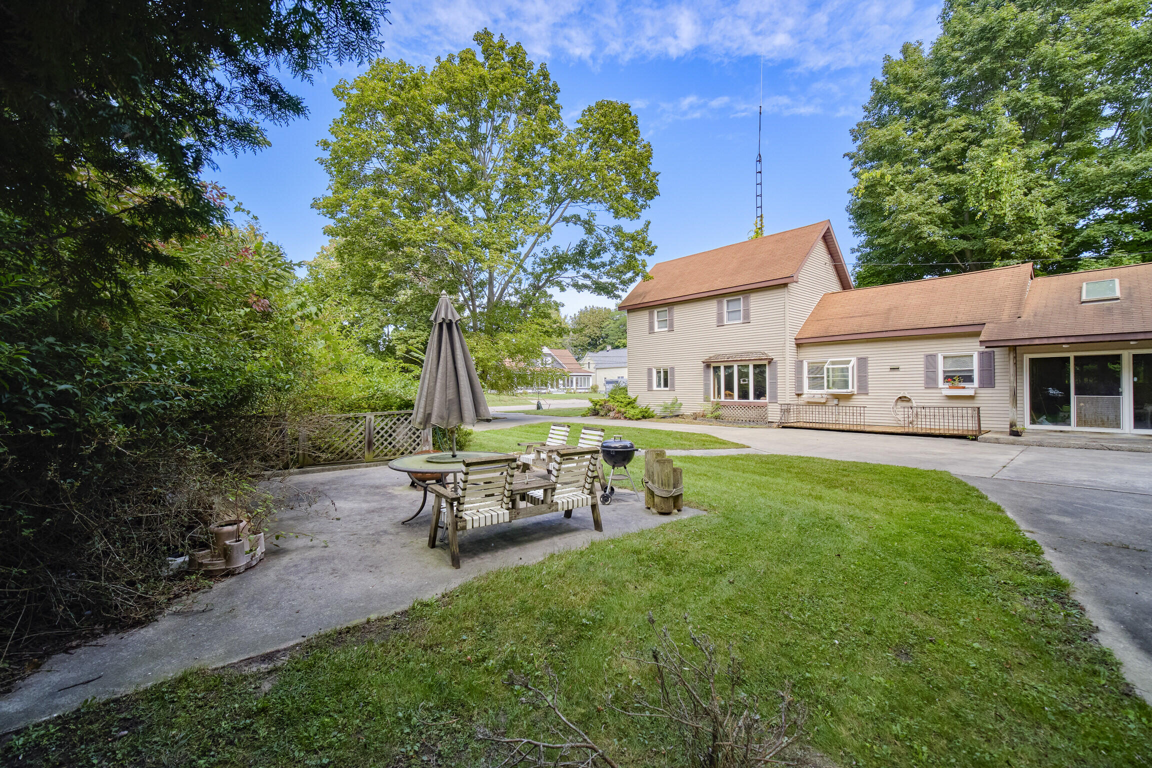 House and green backyard with a paved patio, wooden seating, and barbecue grill.