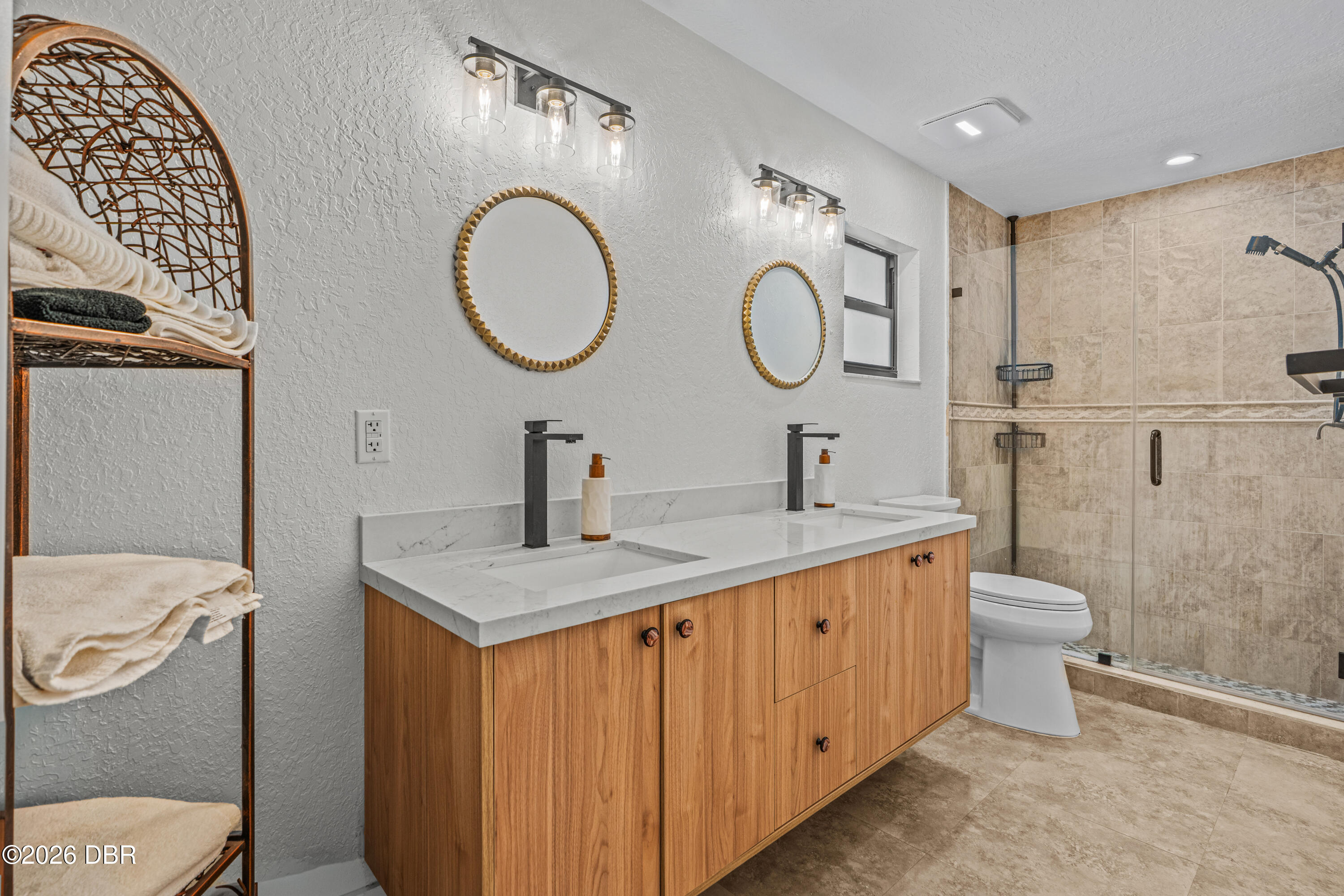 Bathroom with double wood vanity, round gold-framed mirrors, and beige-tiled walk-in shower.